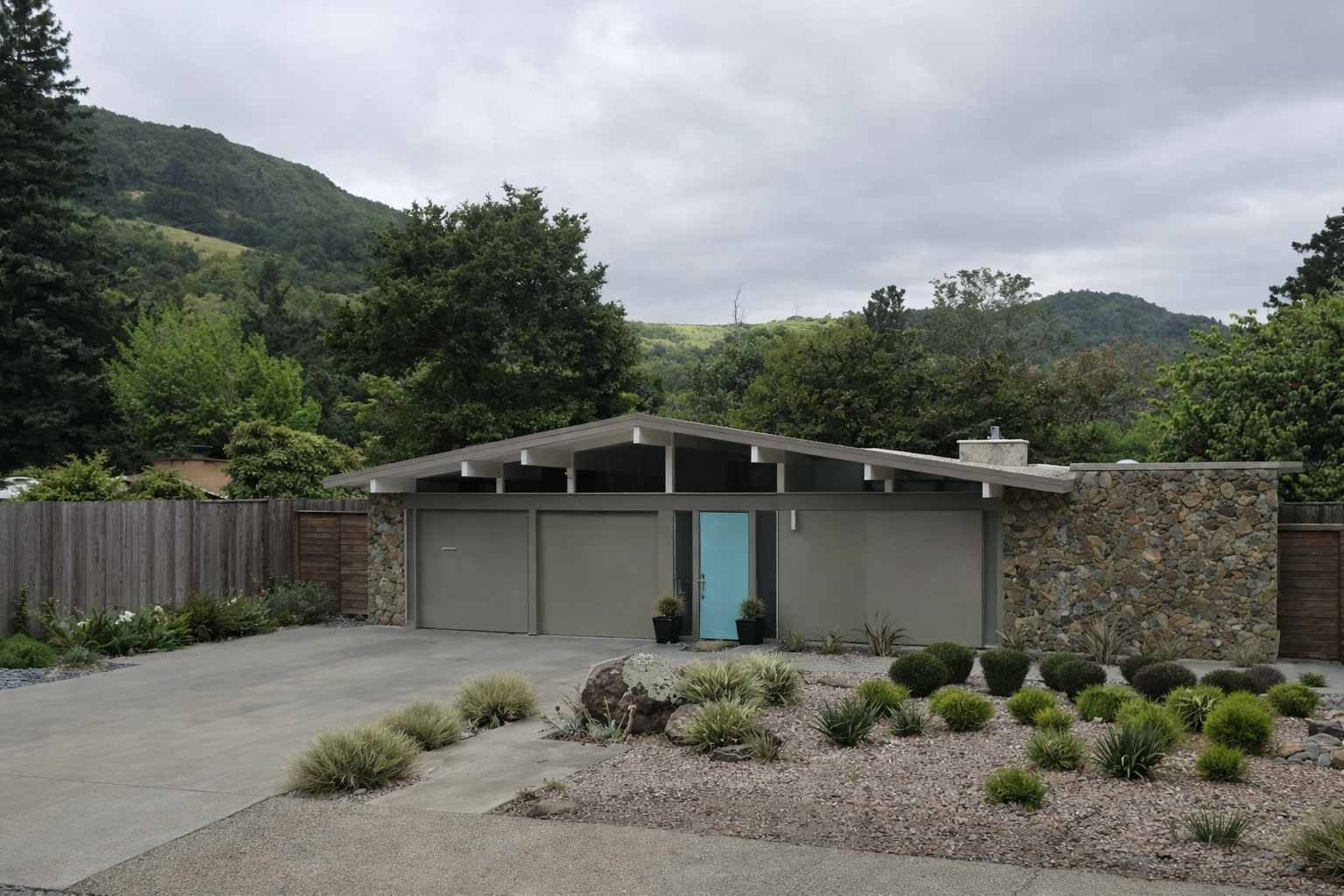 Modern house with gray garage doors, light blue front door, stone and gray exterior, surrounded by desert landscaping, with mountains and cloudy sky in background.