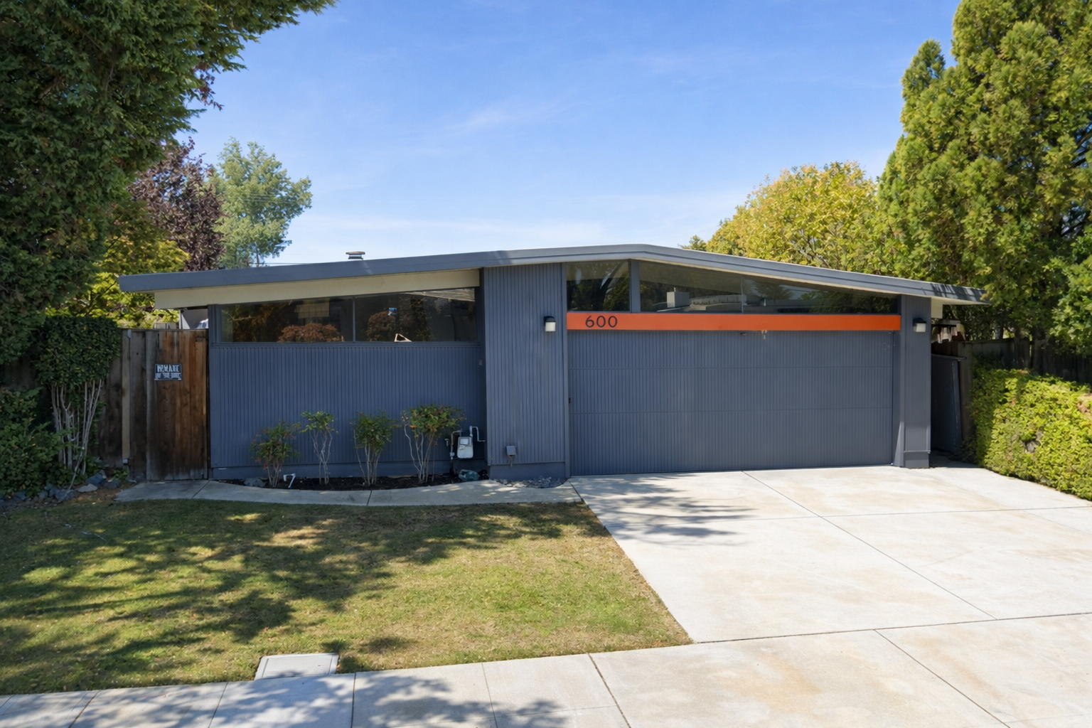 A mid-century modern house with a flat roof, blue exterior, and large window, located on a street with a driveway and some small plants in front.