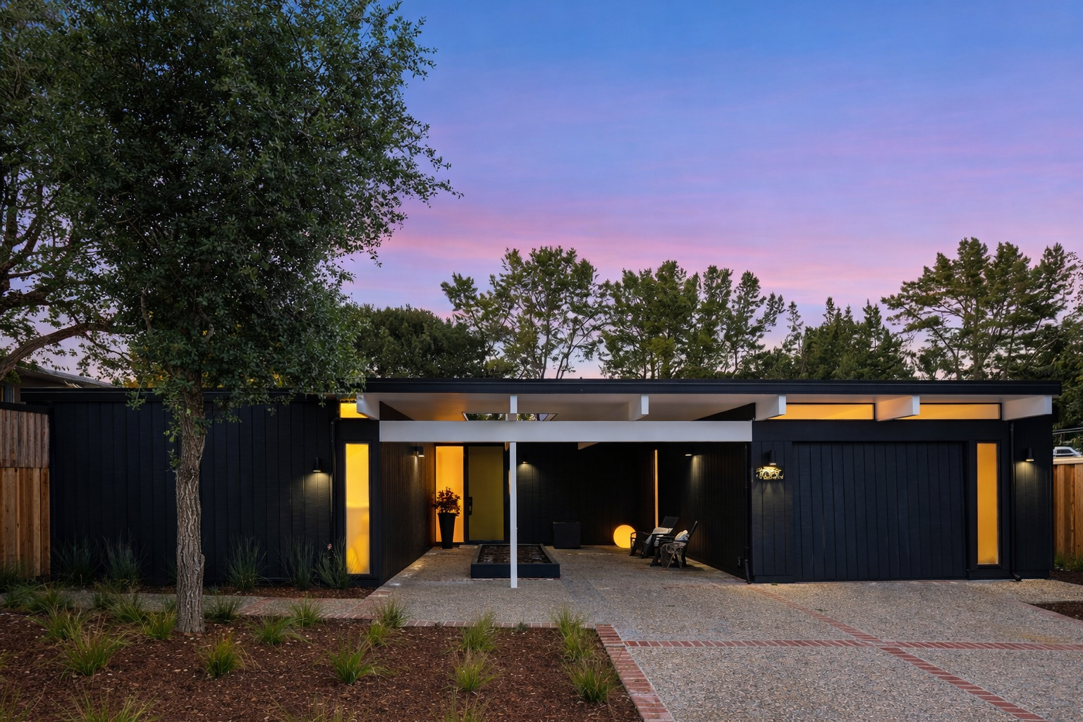 Modern single-story house with black exterior, yellow accents, and a small front patio illuminated by outdoor lights, set against a purple and pink sunset sky with trees in the background.