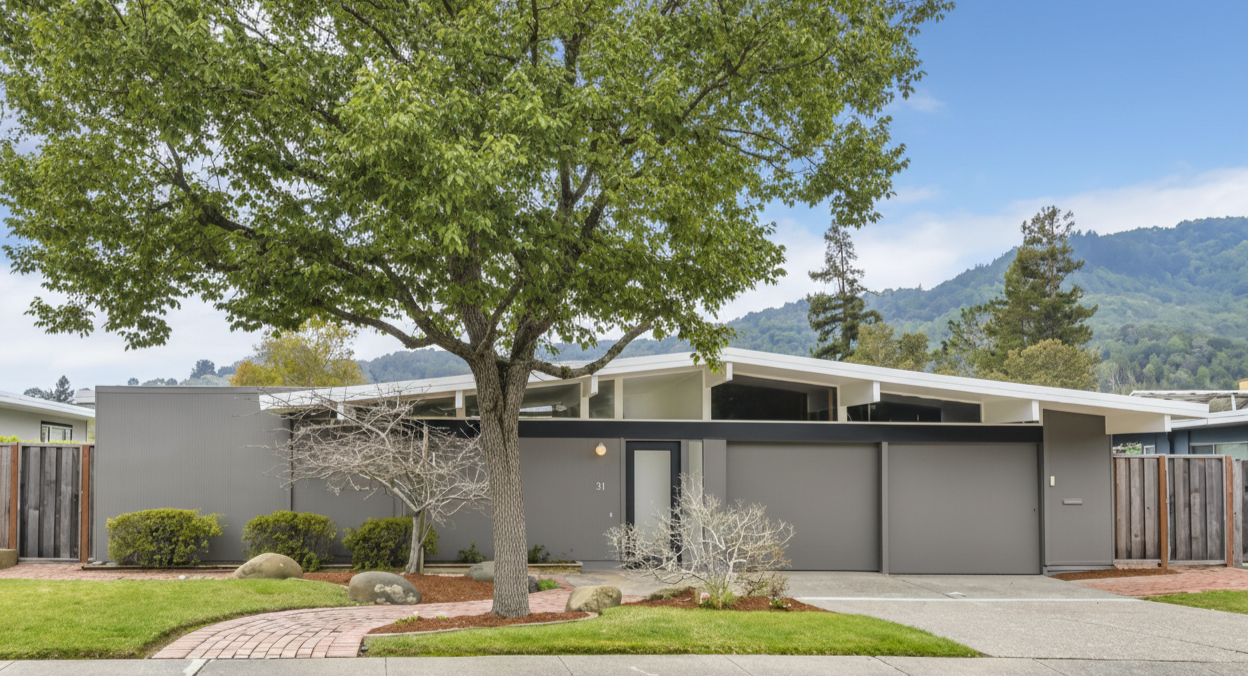 Modern house with gray exterior, large front door, attached garage, landscaped front yard with grass, rocks, and trees, in a setting with mountains and a partly cloudy sky.
