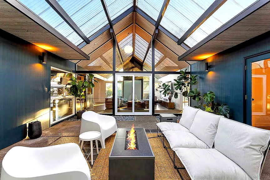 Indoor lounge area with glass ceiling, sofa, chairs, electric fireplace, and potted plants.