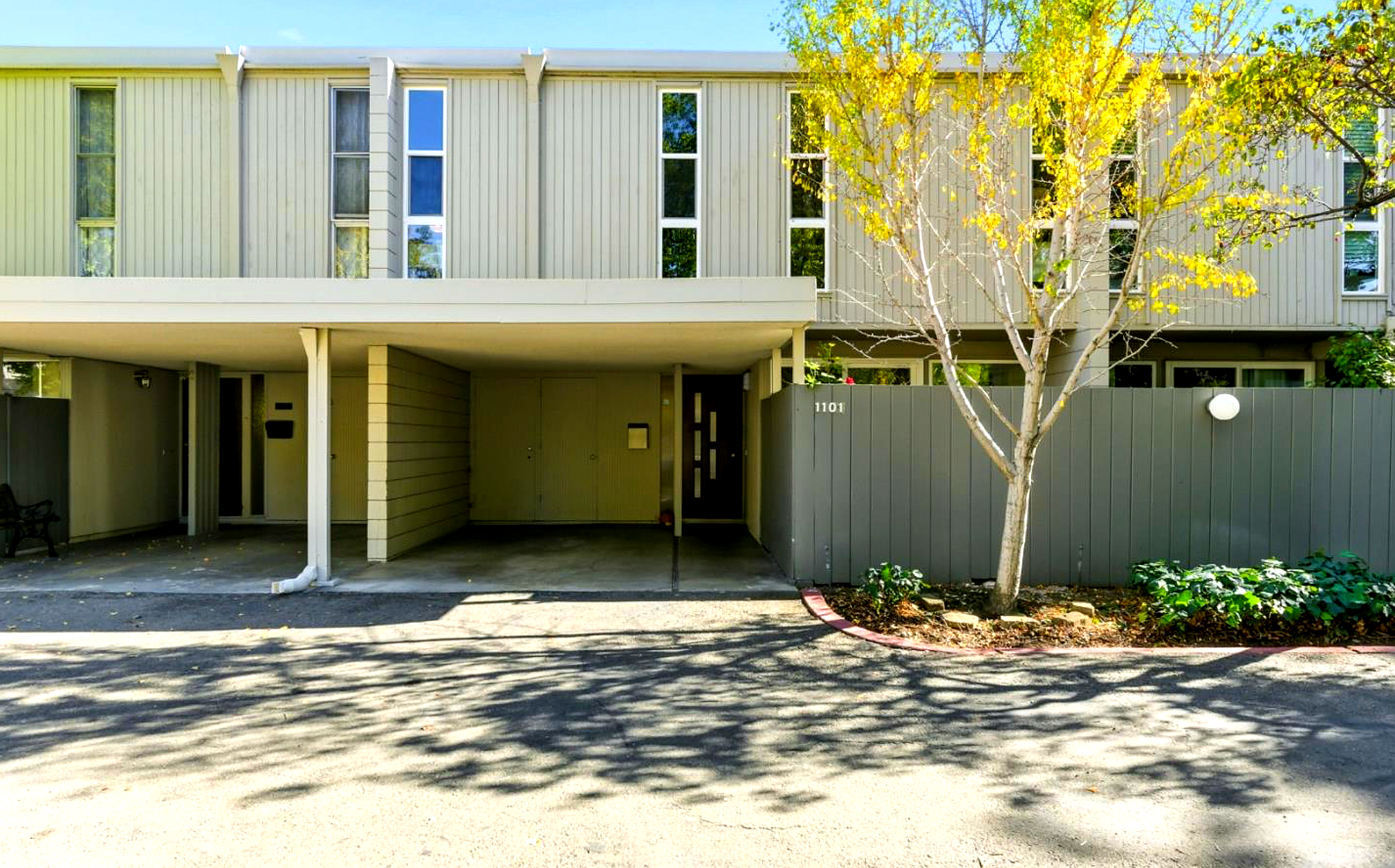 Front view of a modern multi-story apartment building with beige vertical siding and multiple windows, with a tree and landscaped area in front, and an open parking area underneath the building.