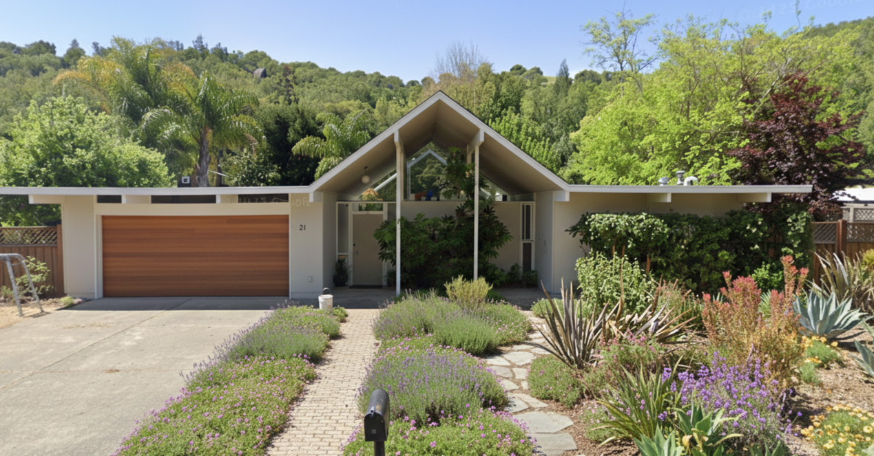 Modern house with triangular roof, surrounded by lush greenery, colorful plants and bushes, with a concrete driveway and a wooden garage door.