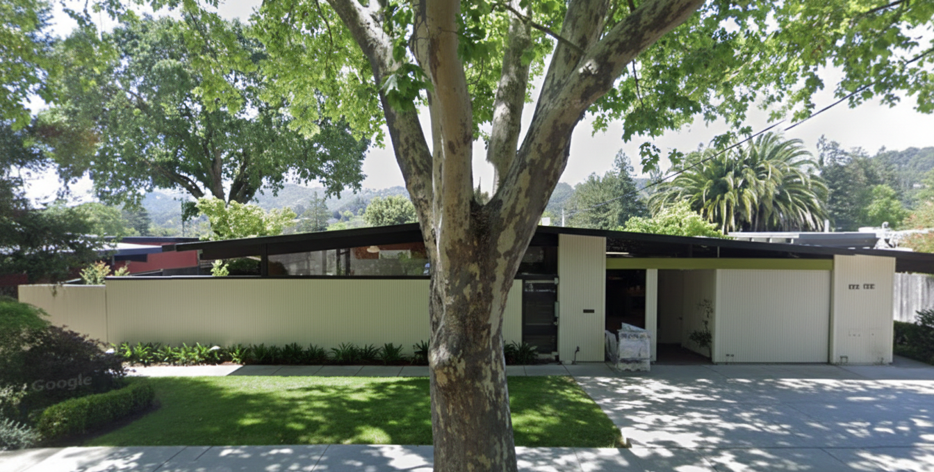 A mid-century modern house with a lawn, large tree in the front yard, and a driveway.