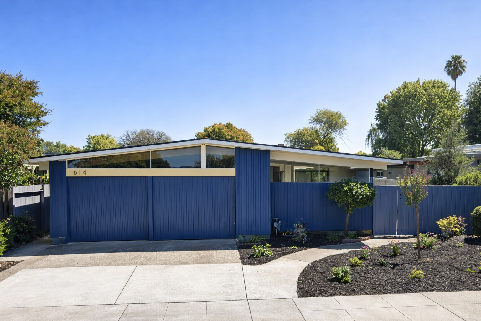 A mid-century modern house with a blue exterior wall and a sloped roof, number 614, surrounded by a front yard with small plants and trees, under a clear blue sky.