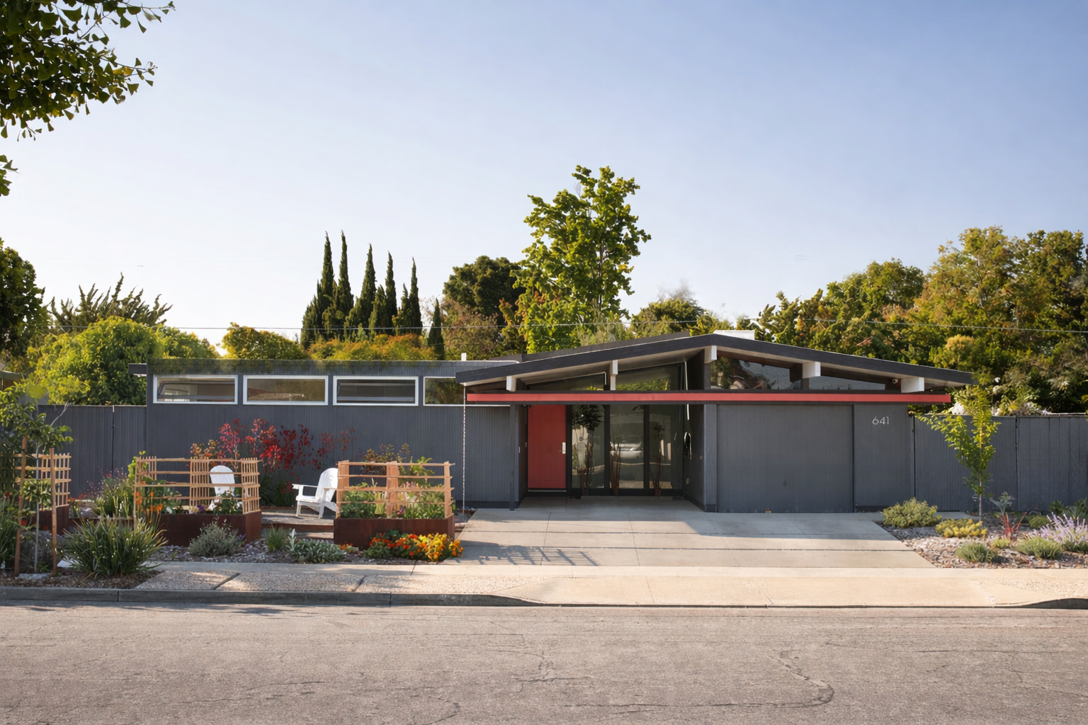 A mid-century modern house with a metal exterior, surrounded by a garden with plants and flowers, has a slightly sloped roof, glass doors, and a sidewalk in front.