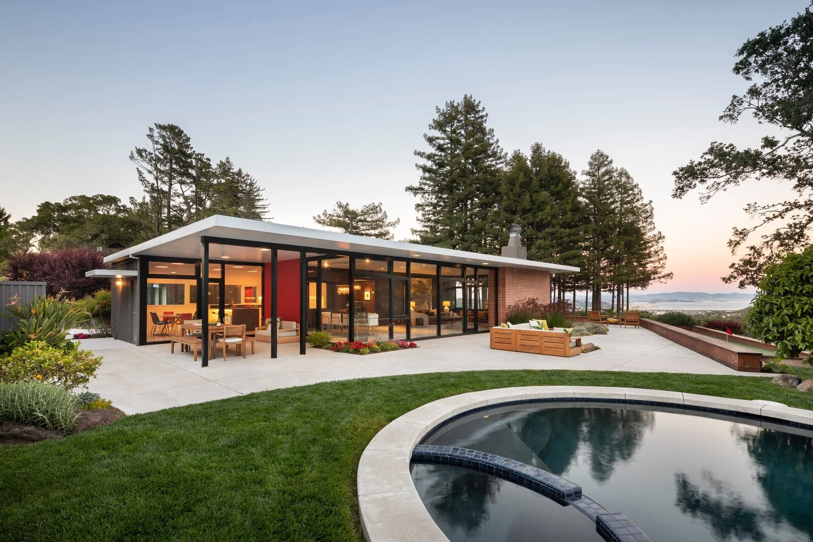 Modern house with large glass windows, outdoor seating area, and a swimming pool in a landscaped yard with trees and a view of a body of water in the distance, during sunset.