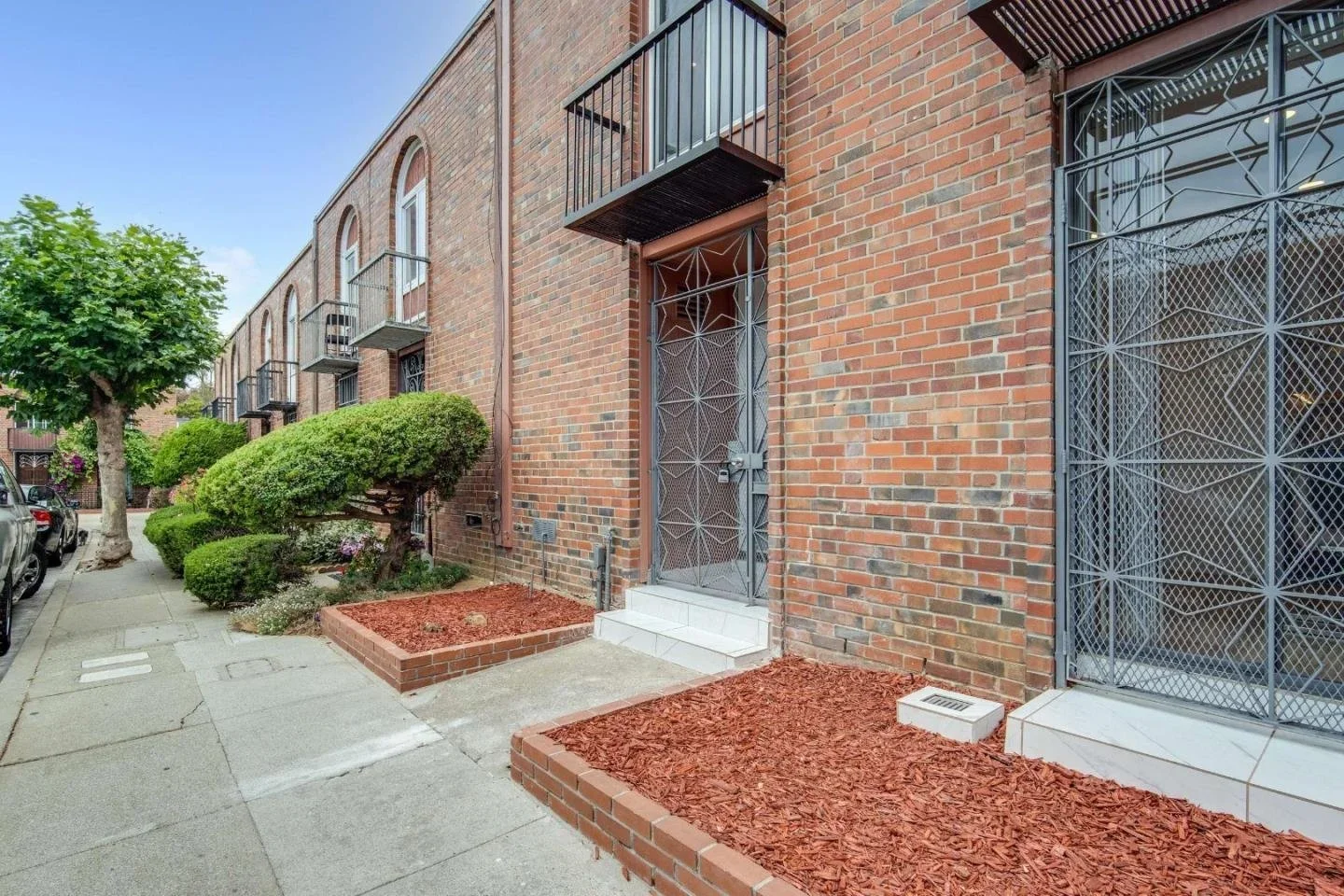 Red brick apartment building exterior with small steps leading to a metal door, window with a metal grille, potted plants, neatly trimmed bushes, and a tree on a sidewalk.