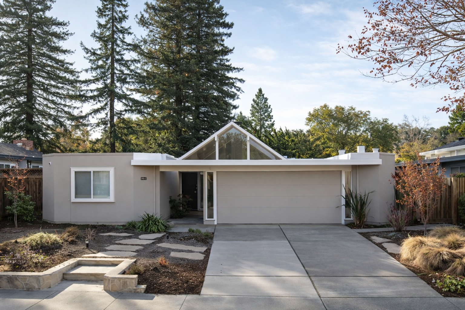 Modern single-story house with a triangular glass front, beige exterior, and a two-car garage, surrounded by trees and landscaped yard.