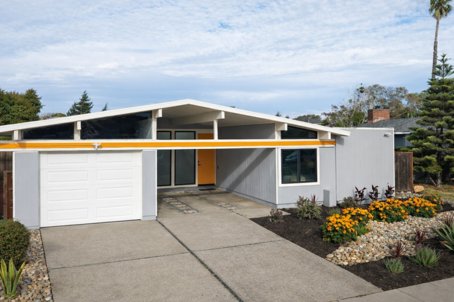 Modern mid-century house with gray exterior, yellow accent door, large front window, and a white garage door. The house features a sloped roof and is surrounded by a landscaped yard with flowers, rocks, and plants.