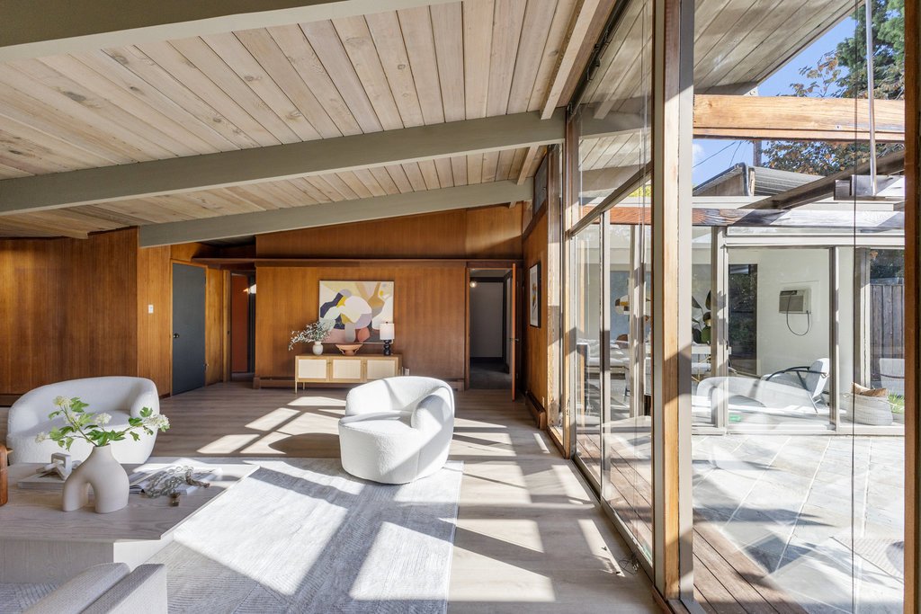 Modern living room with large glass windows, wooden walls, two white curved armchairs, a white coffee table with decorative items, a colorful abstract painting, and an outdoor patio visible through the windows.