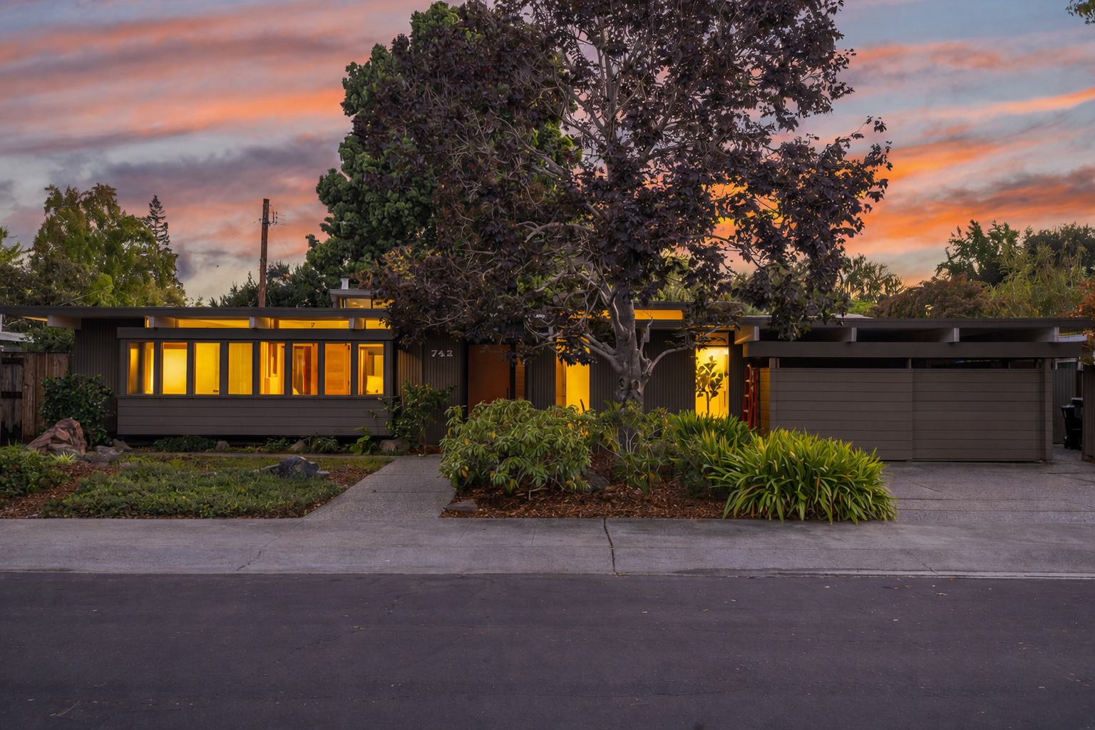 Modern house illuminated from within at dusk with colorful sky, a large tree in front, and a landscaped yard with plants and rocks.
