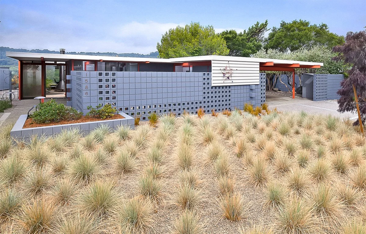 A mid-century modern house with a flat roof and a facade made of grey concrete blocks, surrounded by a landscaped yard with ornamental grasses, shrubs, and trees, under a cloudy sky.