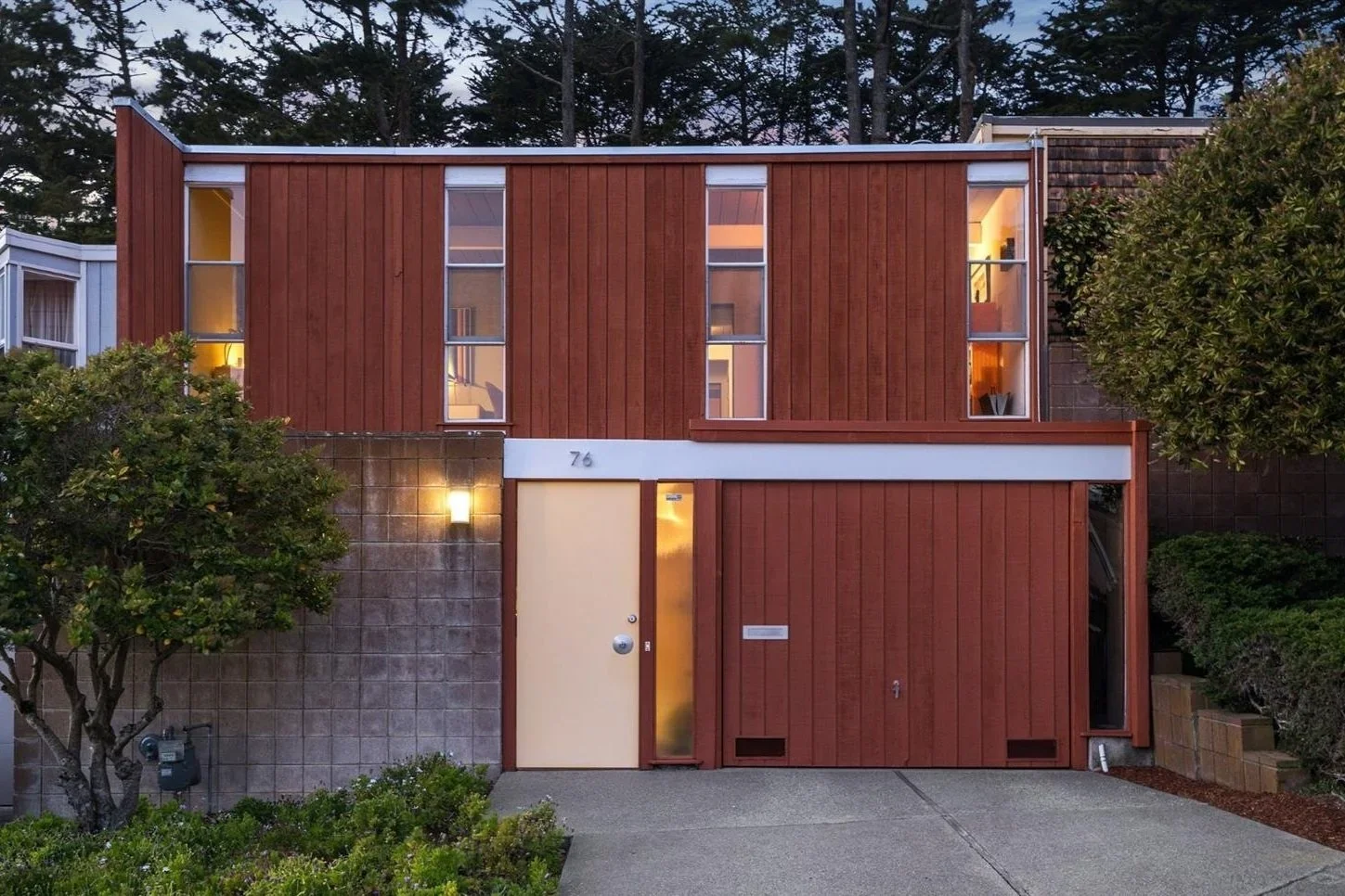 Front view of a modern two-story house with a red wooden exterior, large vertical windows, and a concrete driveway, surrounded by greenery and trees.