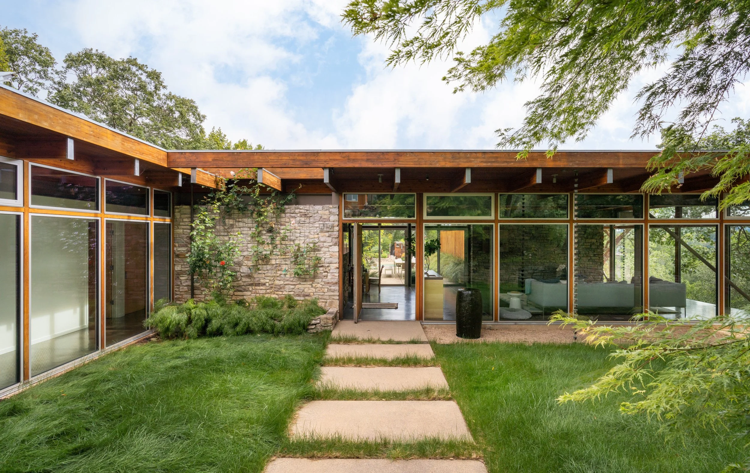 Front view of a modern house with large glass windows, stone wall, wooden roof overhang, a pathway with stepping stones, green lawn, and trees surrounding the house.