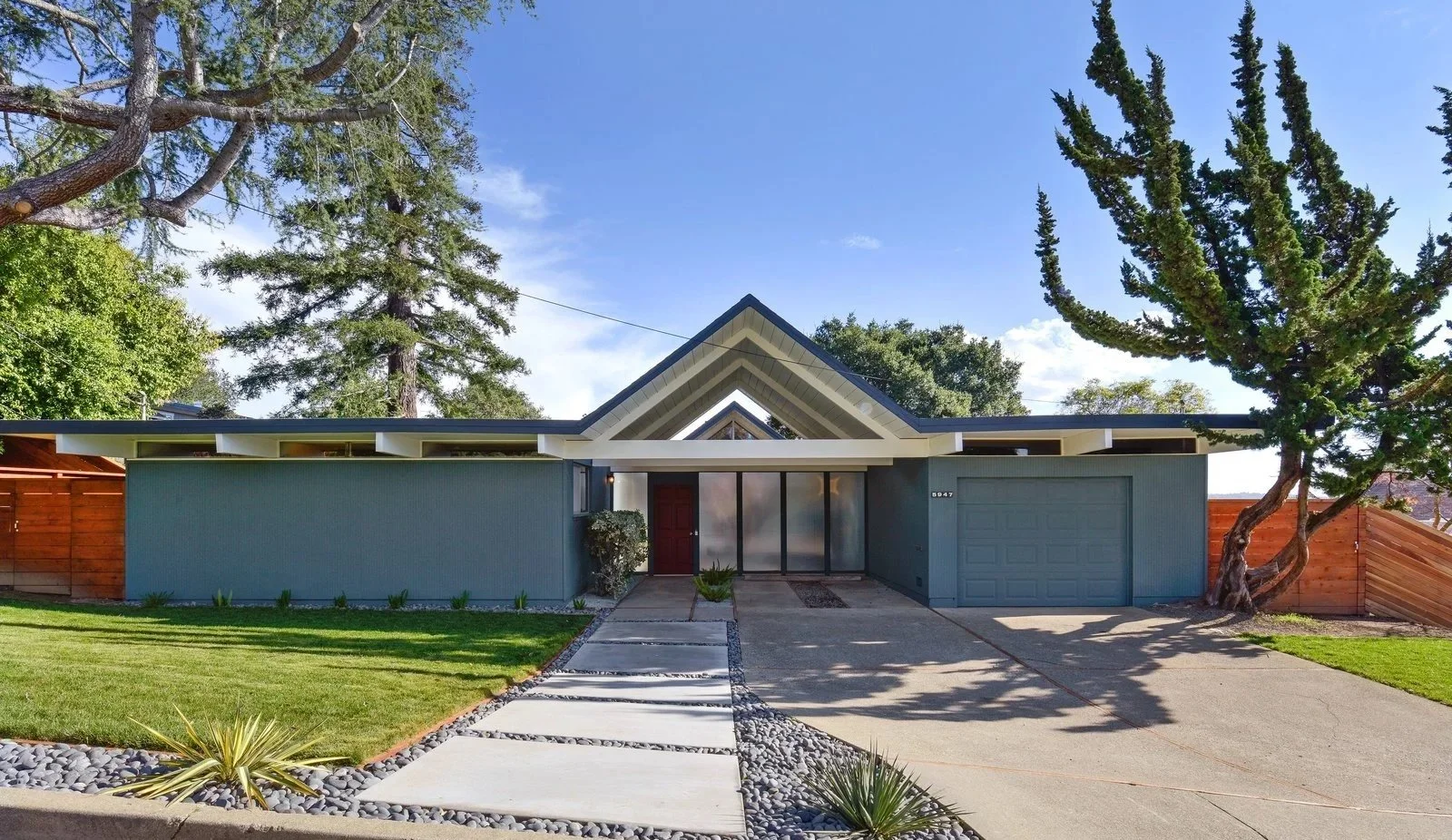 Front view of a mid-century modern house with a gable roof, two large trees on either side, a concrete driveway, and a well-maintained lawn.