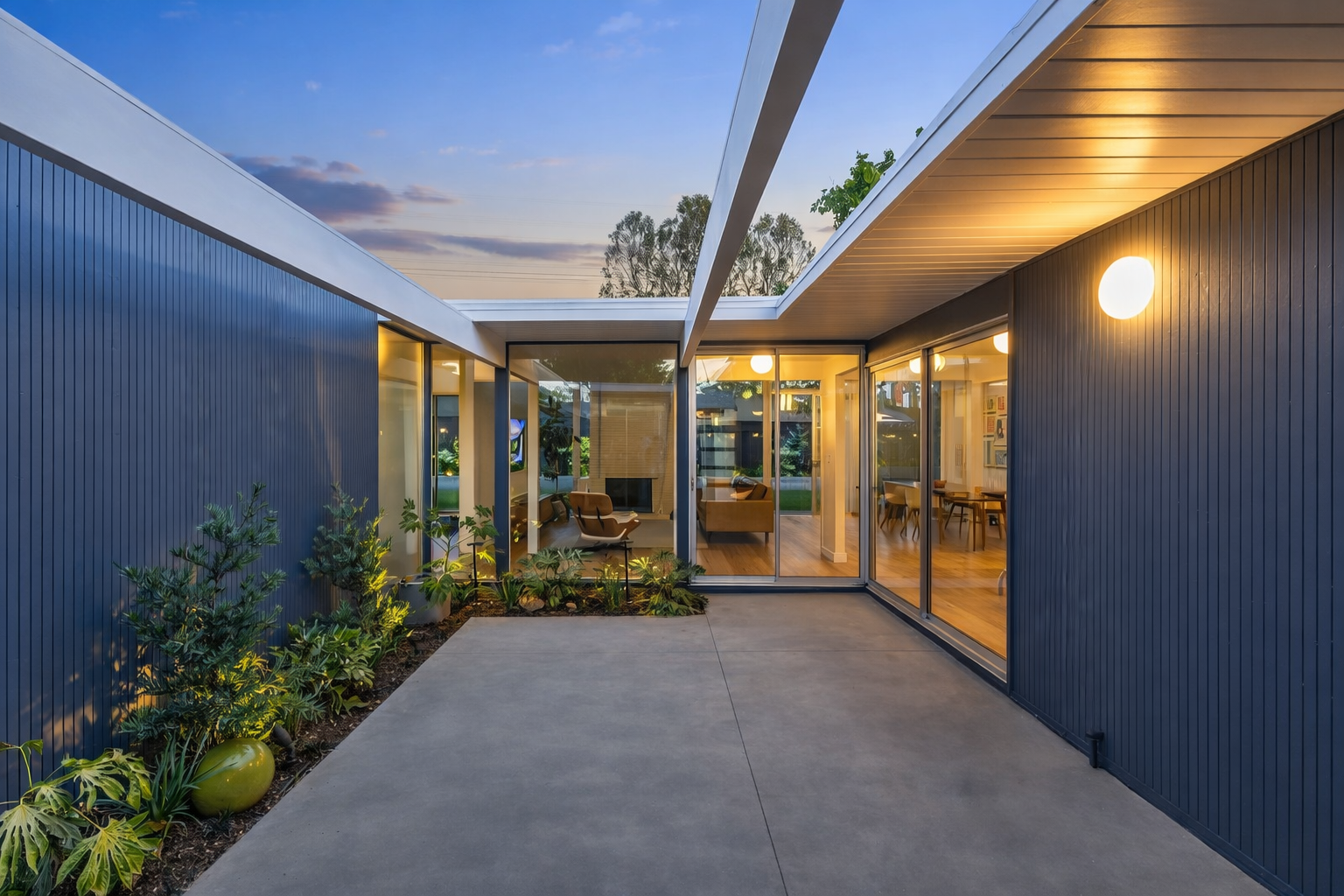 Modern house patio with concrete floor, glass sliding doors, interior with seating area and plants outside, and evening sky with sunset colors.