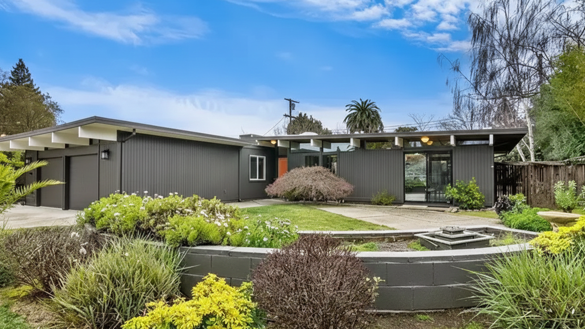 Modern single-story house with black siding, large windows, and surrounding landscaped garden with various shrubs and plants under a partly cloudy sky.