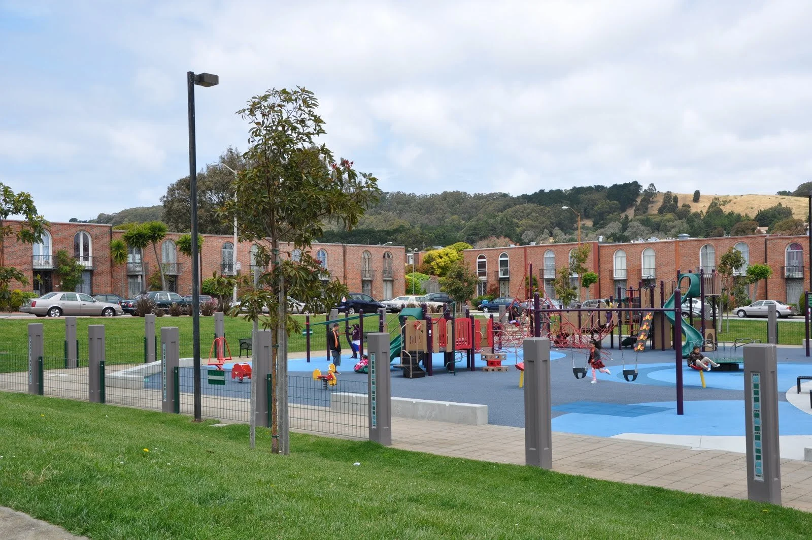 A colorful playground with slides, swings, and climbing structures, surrounded by a fence, in a residential area with parked cars and brick apartment buildings in the background, under a partly cloudy sky.