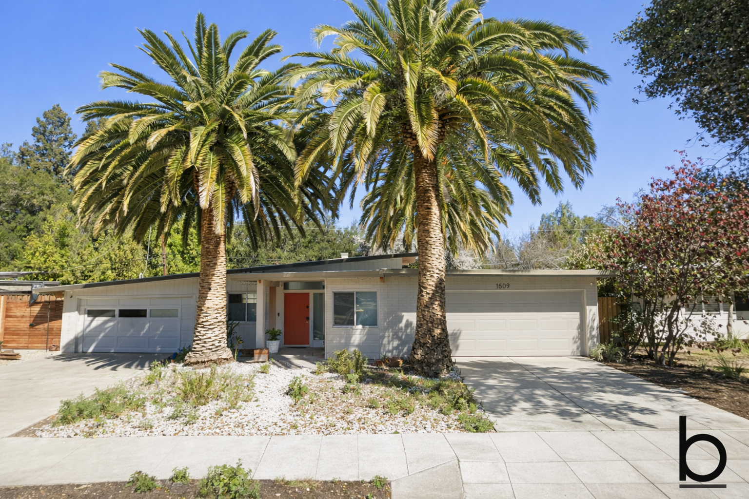 Front view of a mid-century modern house with a flat roof, white brick exterior, and a red front door, flanked by two large palm trees.