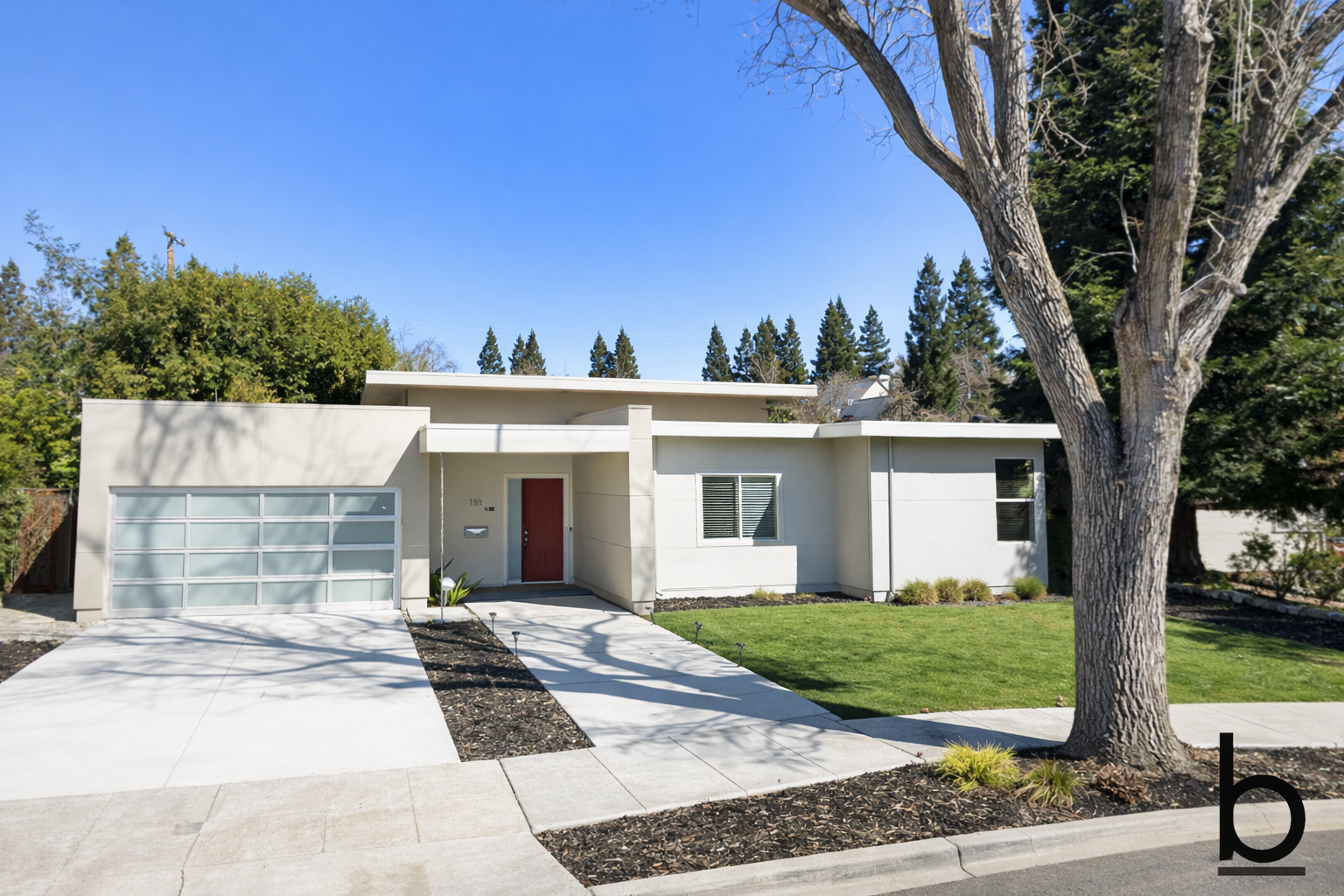 Modern white house with a red front door, a large garage door with glass panels, a well-maintained lawn, and a large tree in front under a clear blue sky.