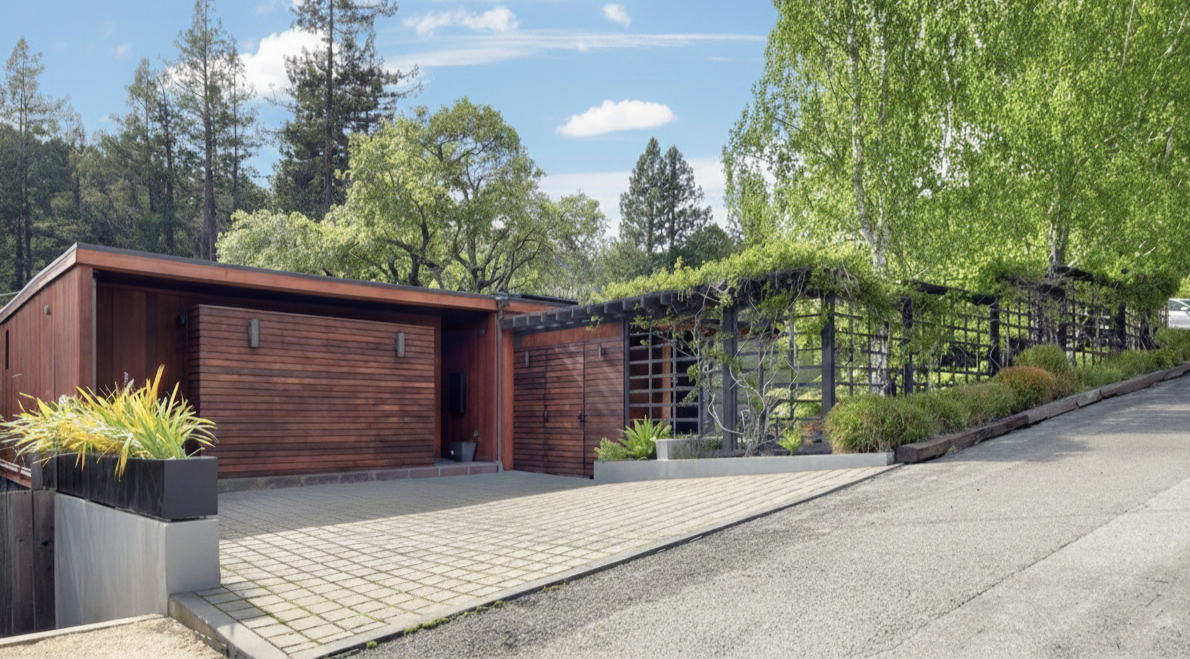 Modern house with wooden exterior, surrounded by trees and greenery, with paved driveway and garden plants.