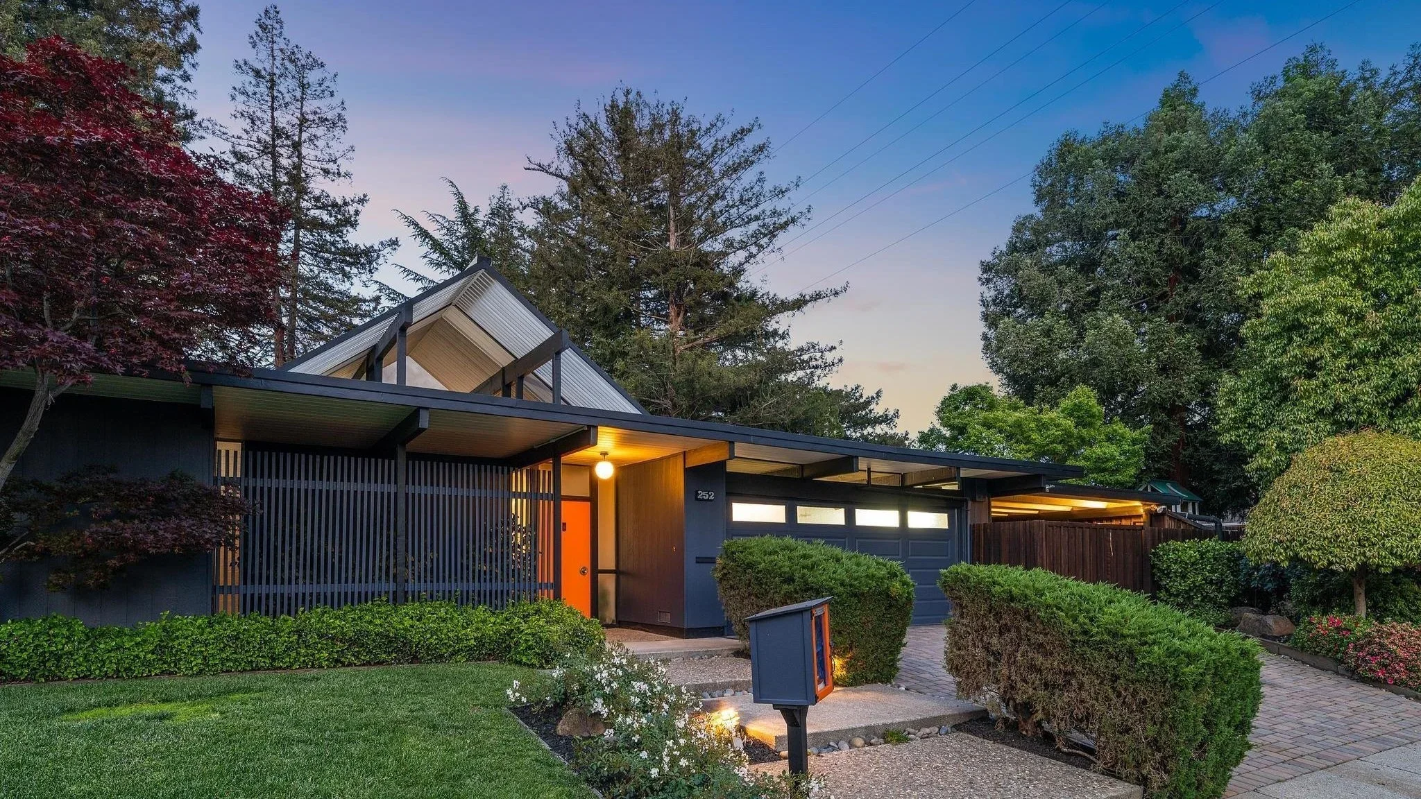 Modern house with dark exterior, orange front door, garage, surrounded by green bushes, trees, and a well-maintained lawn, evening with lights on.