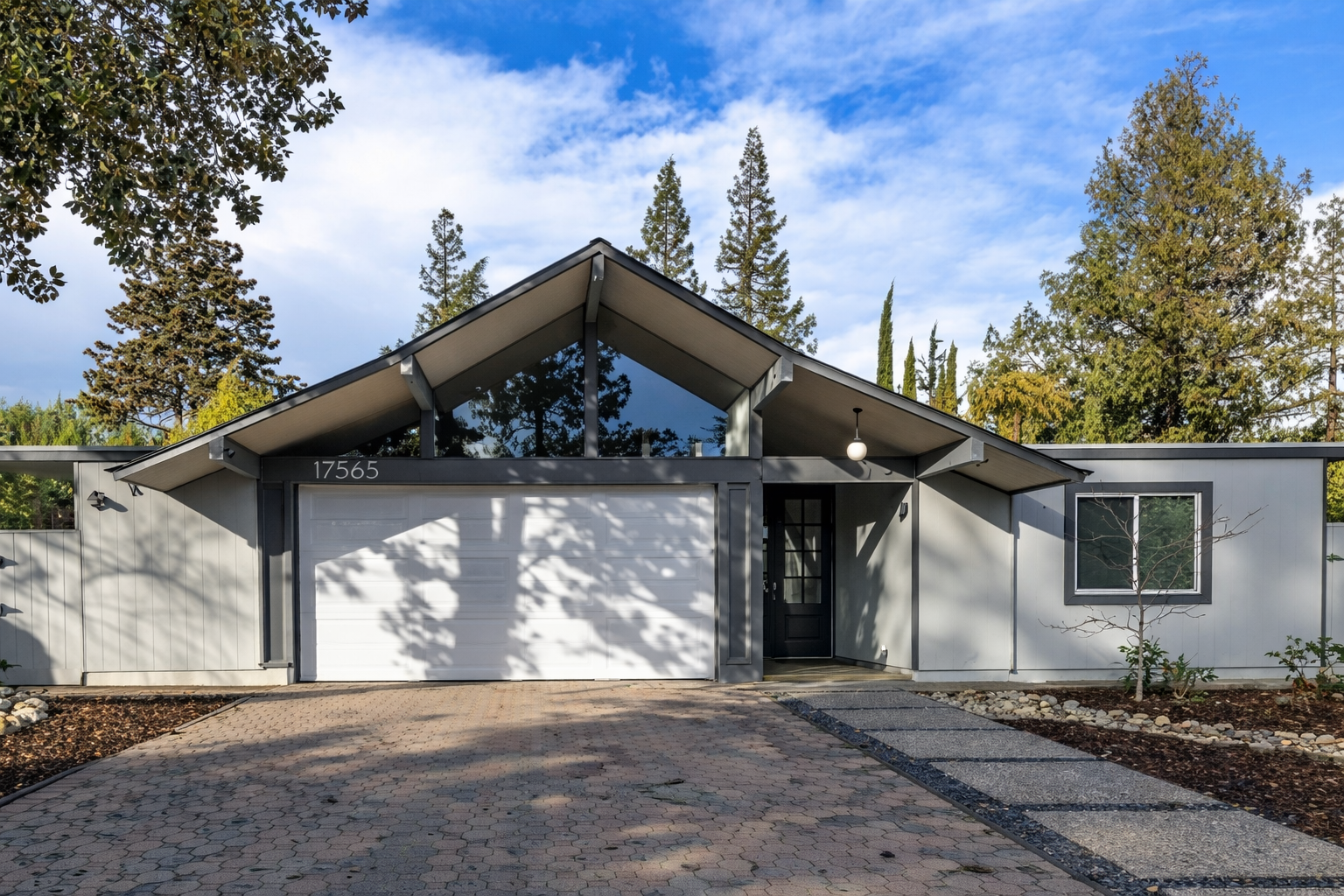Modern house with white and black exterior, garage door, and a front yard with a pathway and small trees, surrounded by tall trees under a blue sky with clouds.