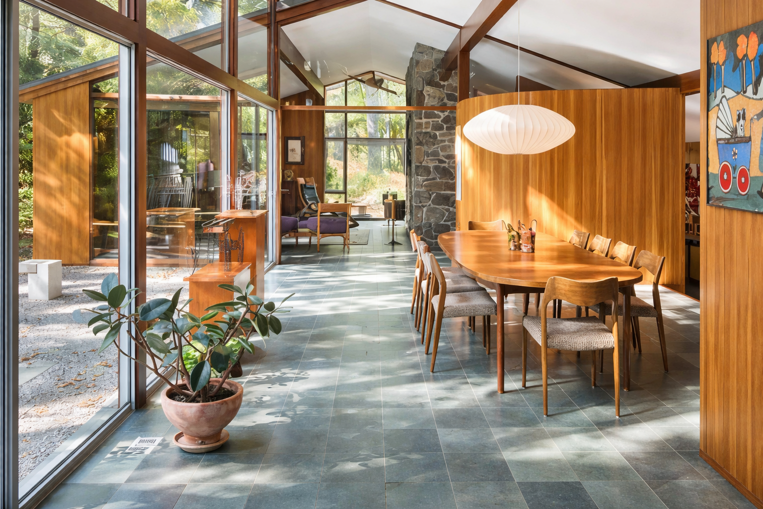 Sunlit interior of a mid-century modern home with wooden paneling, large windows, and a green tiled floor. Features a wooden dining table with chairs, a potted plant, and artwork on the walls.