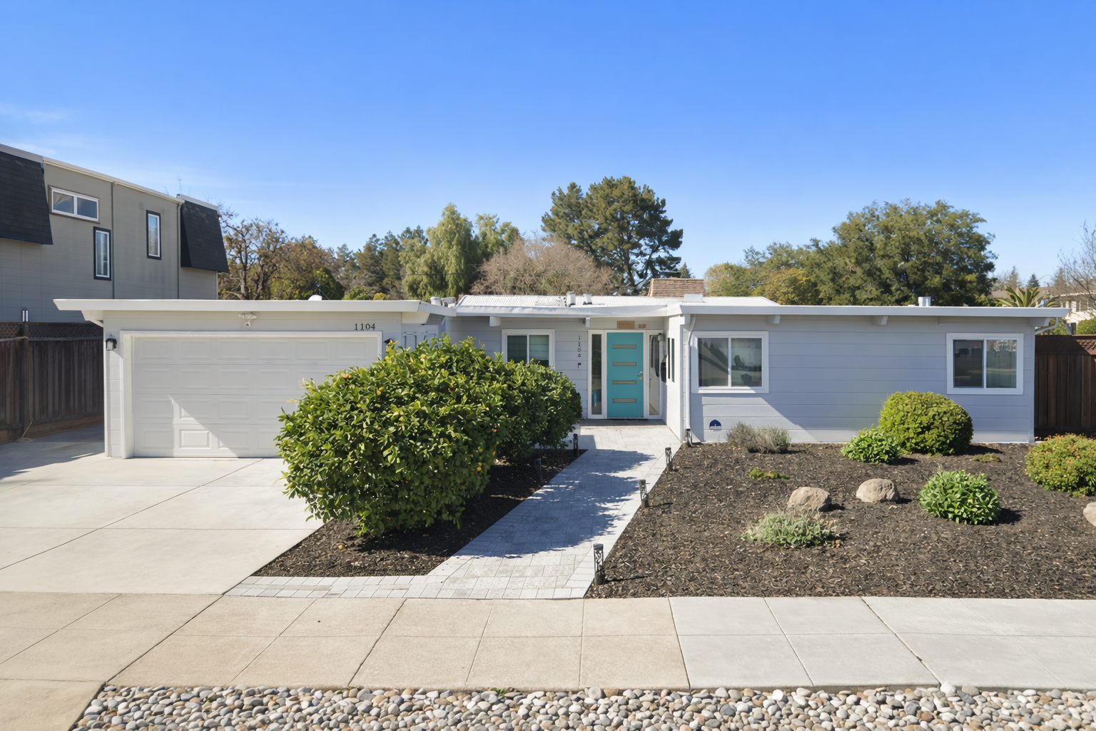 Front view of a modern single-story house with a light blue door, white exterior, surrounding landscaped garden, and a concrete driveway.