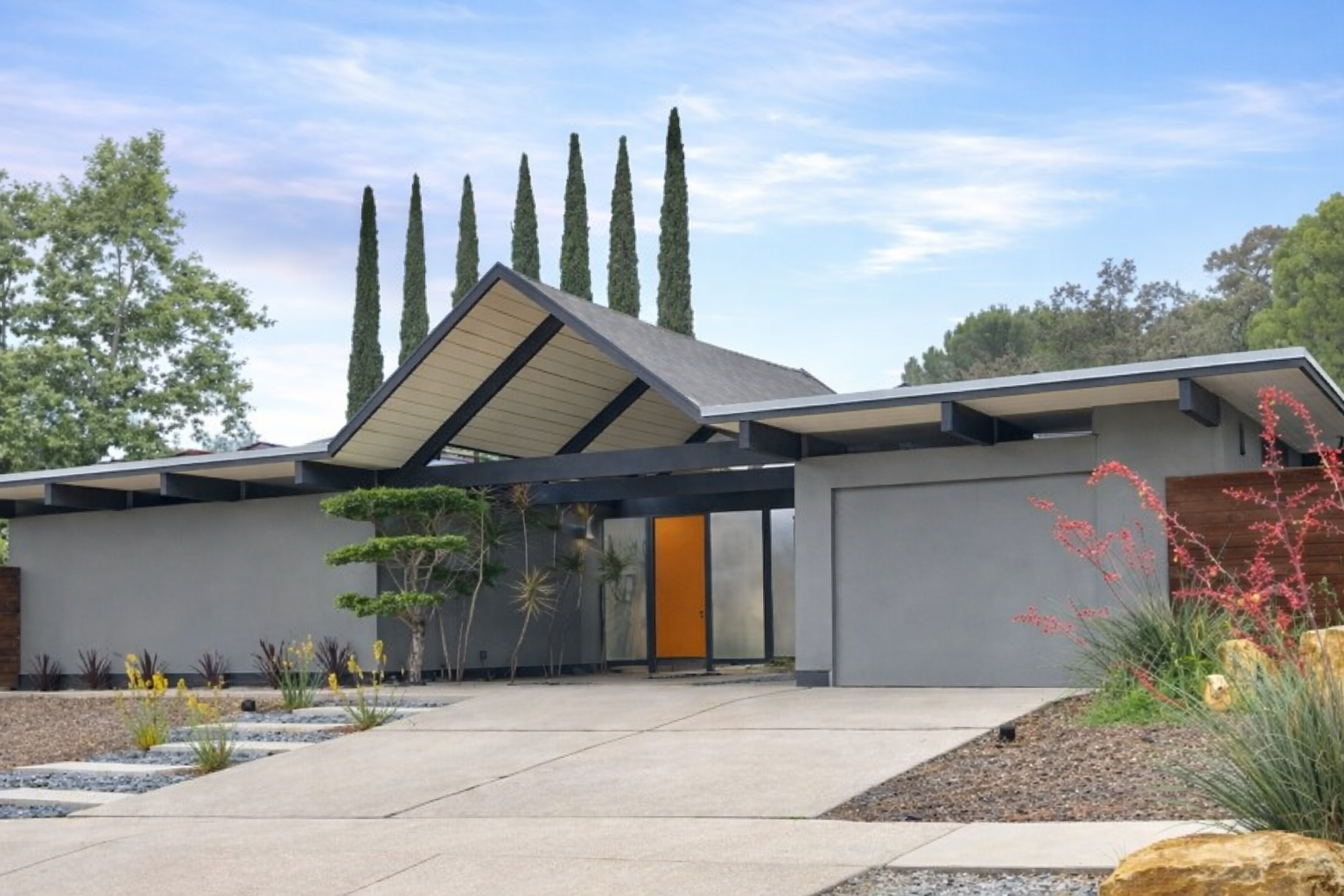 Front view of a modern residential house with a flat roof, gray exterior walls, and an orange front door. Landscaping includes small trees, drought-tolerant plants, and rocks, with tall cypress trees in the background under a partly cloudy sky.
