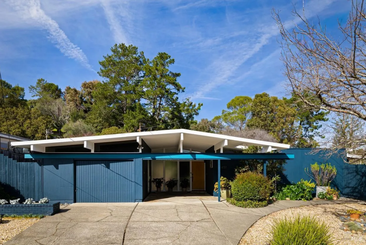 Front view of a mid-century modern house with a flat roof, blue exterior walls, and a concrete driveway, surrounded by trees and gardens.