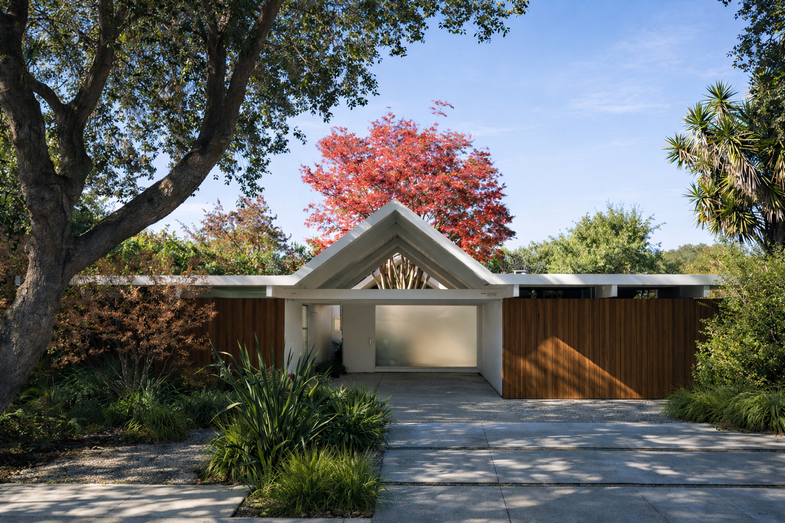 Modern house with a white triangular roof and wooden fence, surrounded by lush greenery and trees with fall foliage, under a clear blue sky.