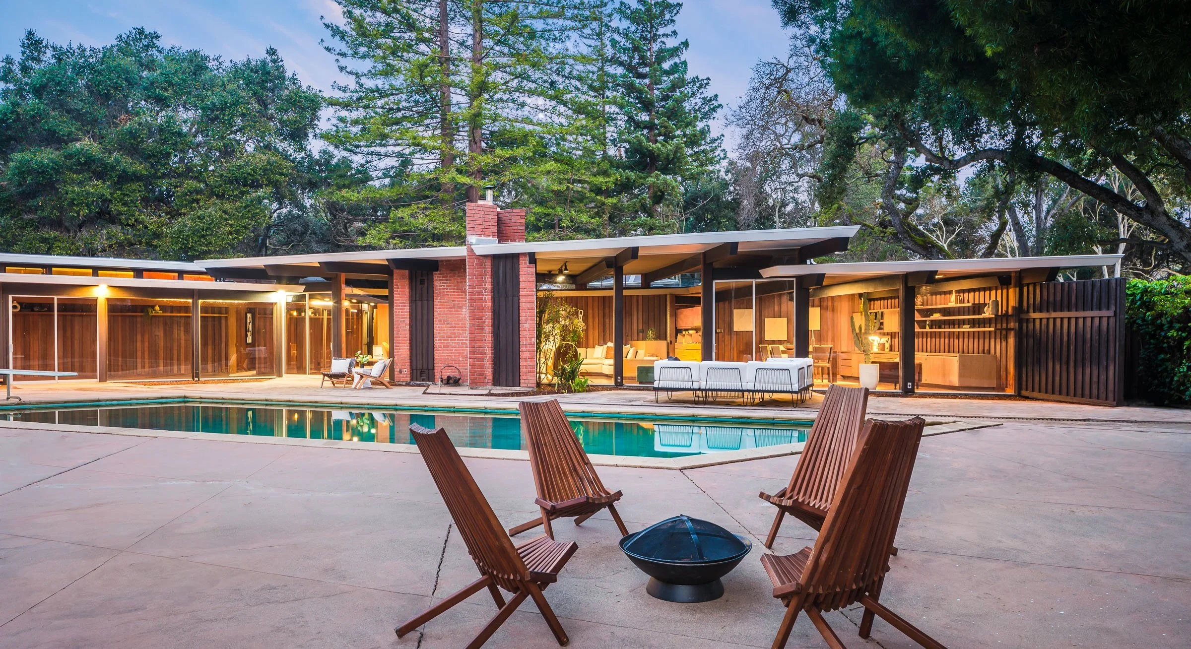 Modern house with large glass windows and an outdoor pool, surrounded by trees, with outdoor furniture and a fire pit on the patio.