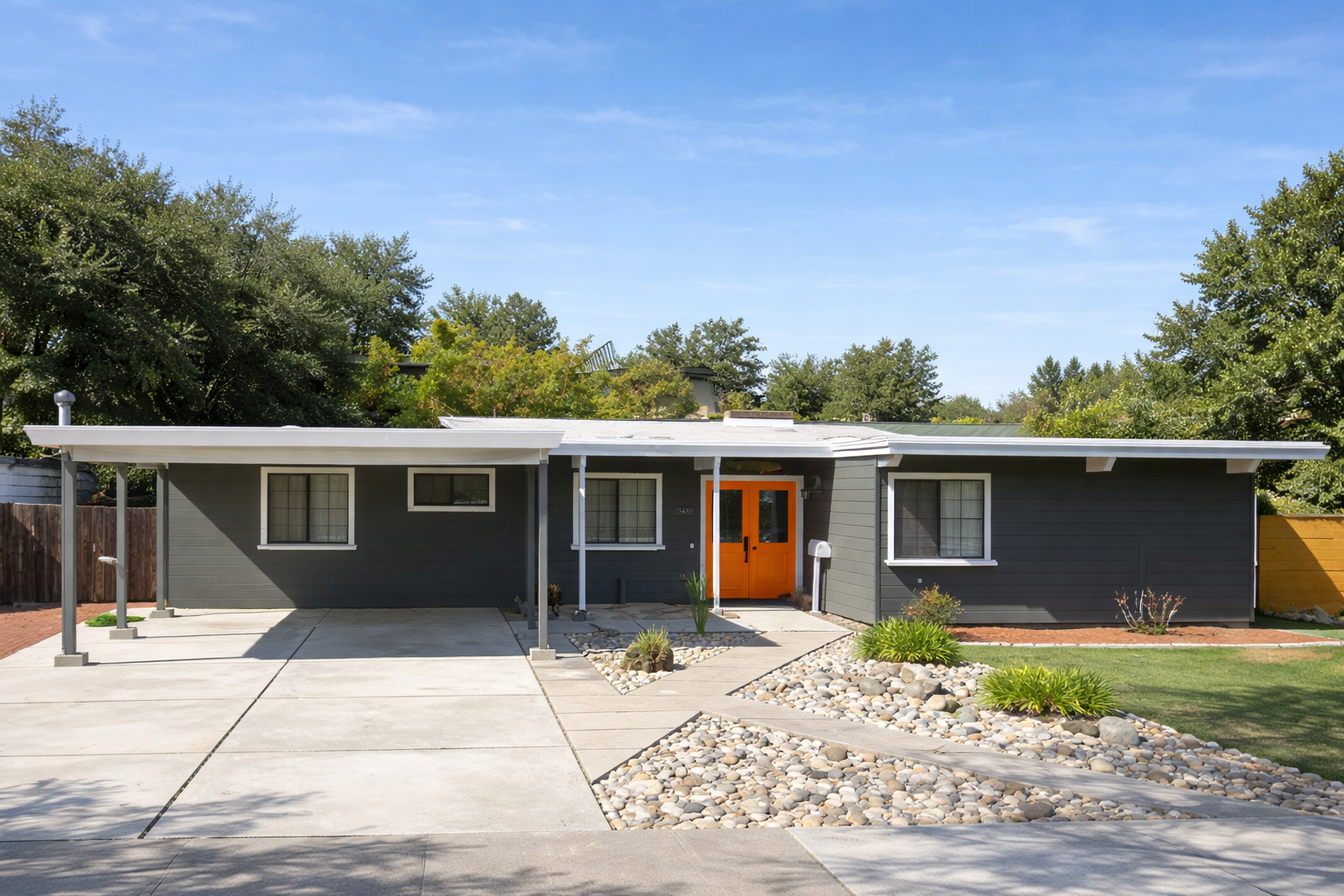 Front view of a modern single-story house with a dark gray exterior, orange front door, and a concrete driveway with a landscaped yard featuring rocks and small plants.