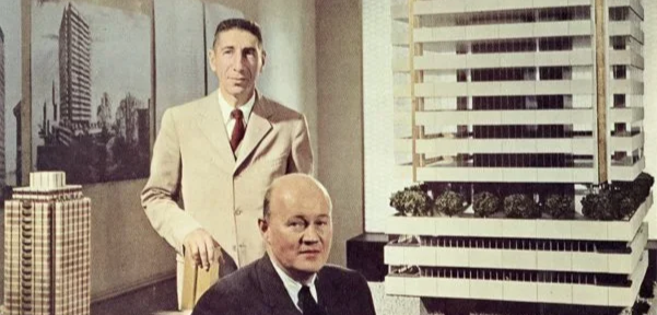 Two men in a room with large buildings in the background, one standing and one sitting.