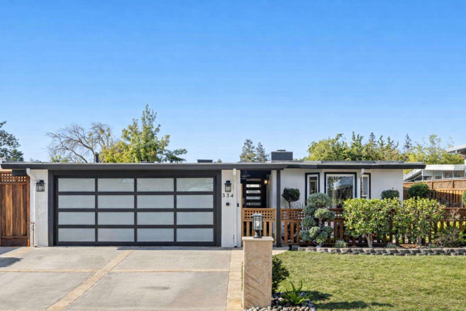 Front view of a modern single-story house with a gray garage door, a small front porch, and landscaped yard with shrubs and plants under a clear blue sky.