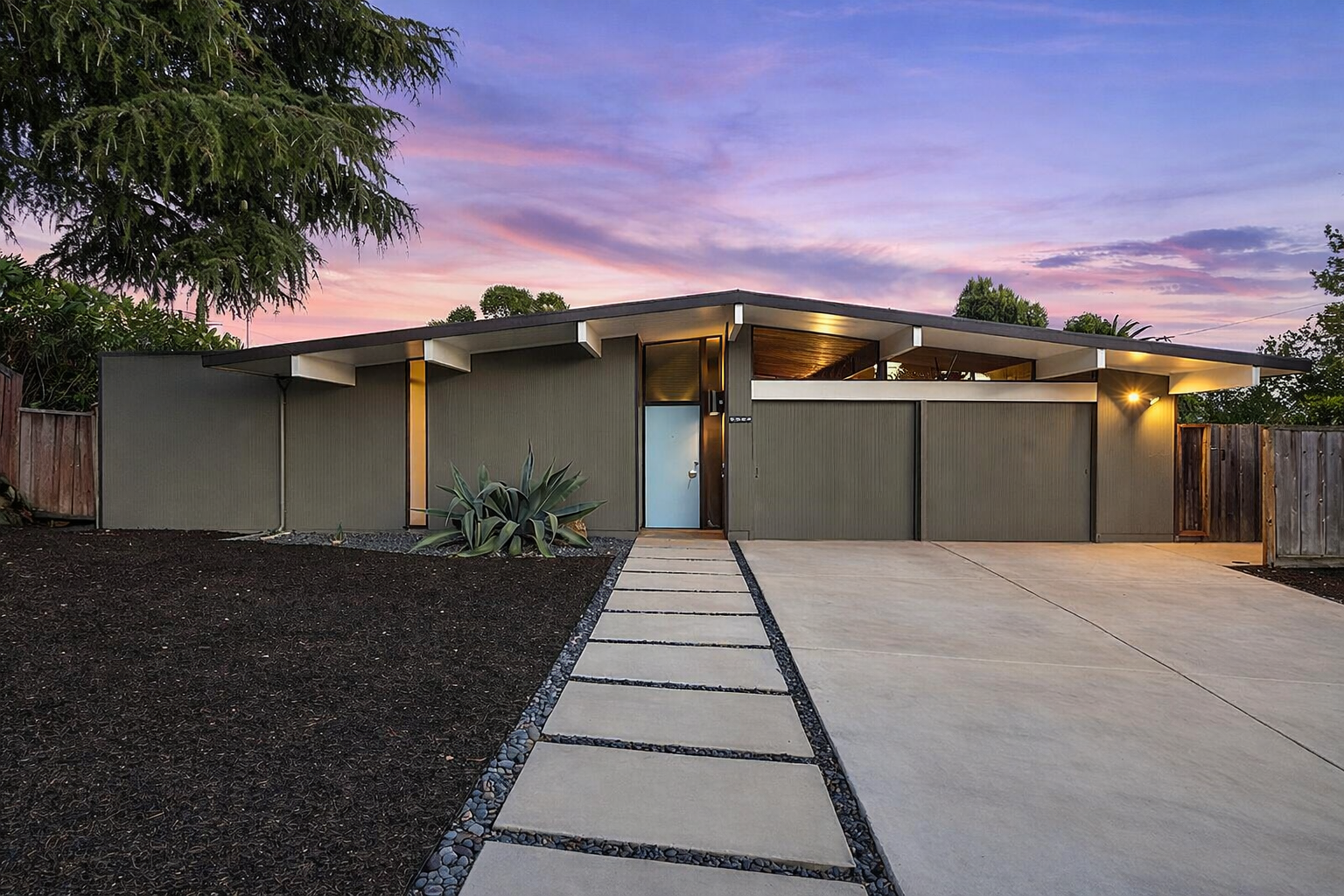 Modern single-story house with a flat roof, landscaped front yard, concrete walkway, and wooden fencing at sunset.