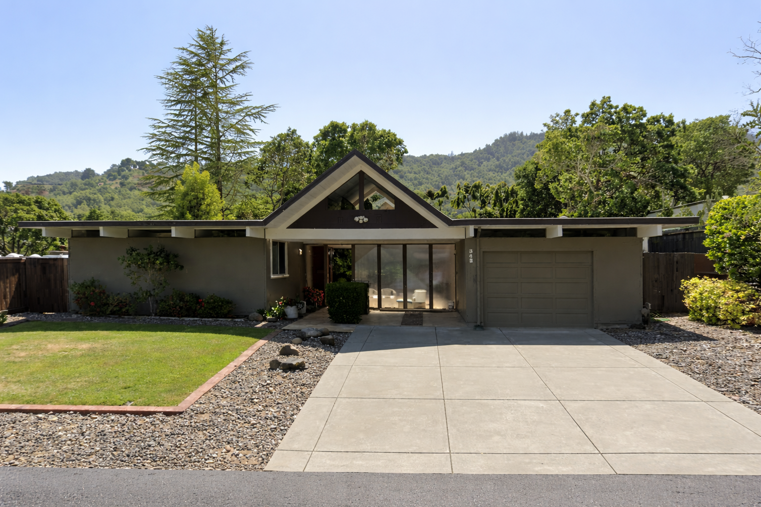A modern, single-story house with a front driveway, a garage on the right, and a glass front entrance in the center, surrounded by greenery and trees.