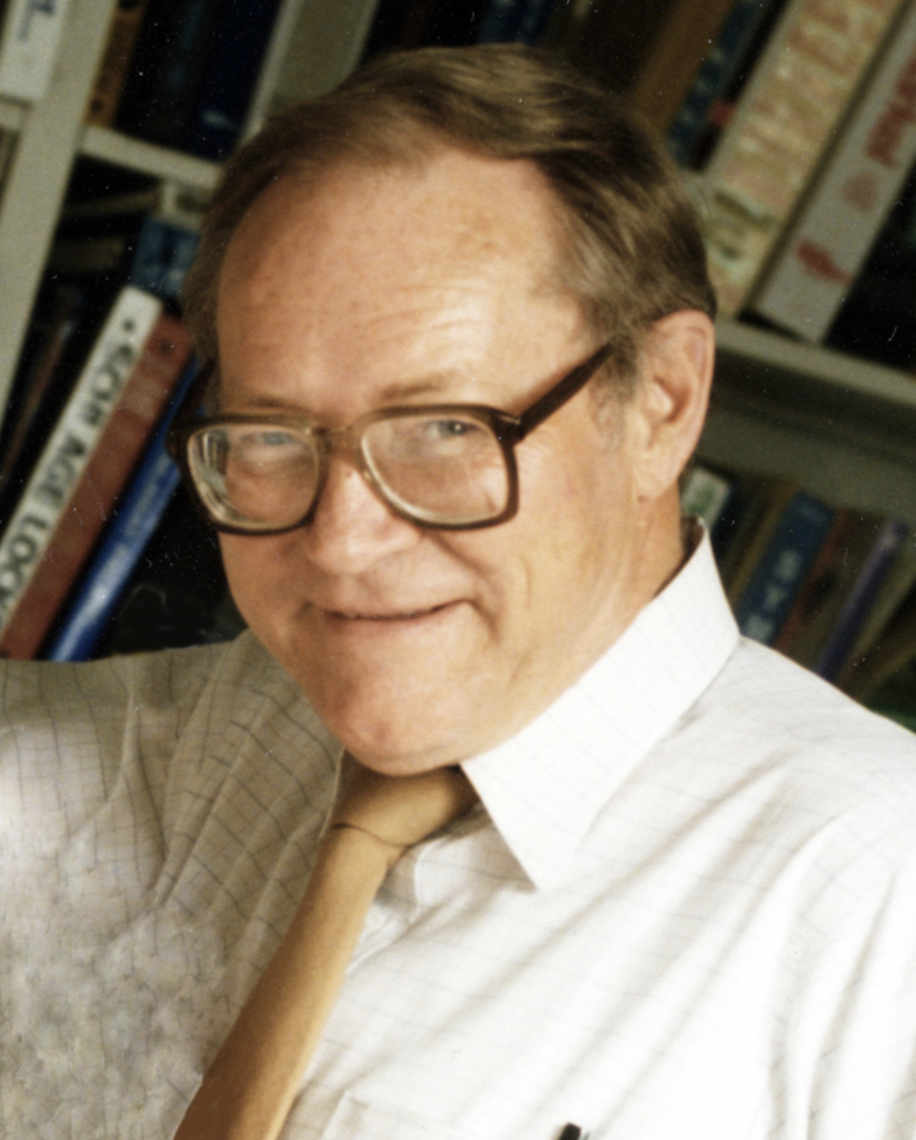 A middle-aged man with light brown hair, wearing glasses, a white checked shirt, and a beige tie, smiling in front of a bookshelf filled with books.