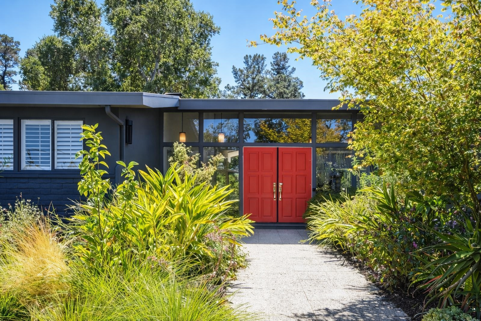 Modern house entrance with a red double door, surrounded by lush green plants and trees, under clear blue sky.
