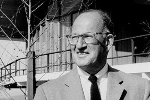 Black and white photo of a man in a suit and tie wearing glasses, outdoors near a power pole and building.