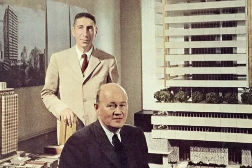 Two men in formal suits in an office with model buildings and cityscape pictures on the wall, and window blinds in the background.
