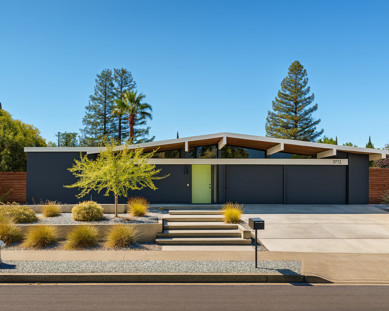 Modern single-story house with dark blue exterior, light green front door, and large front windows. Yard with desert landscaping, including small bushes, a tree, and a few plants. Clear blue sky in the background.