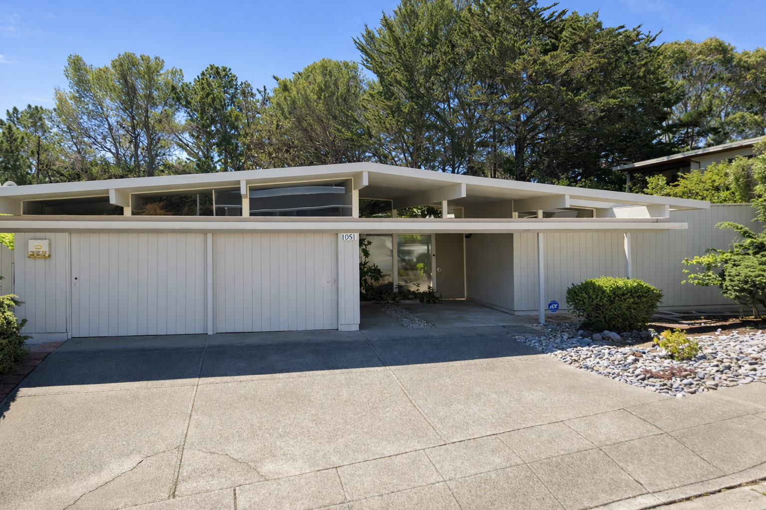 Mid-century modern house with white exterior siding, flat roof with overhangs, garage door, glass entrance door, and landscaped yard with bushes and rocks.