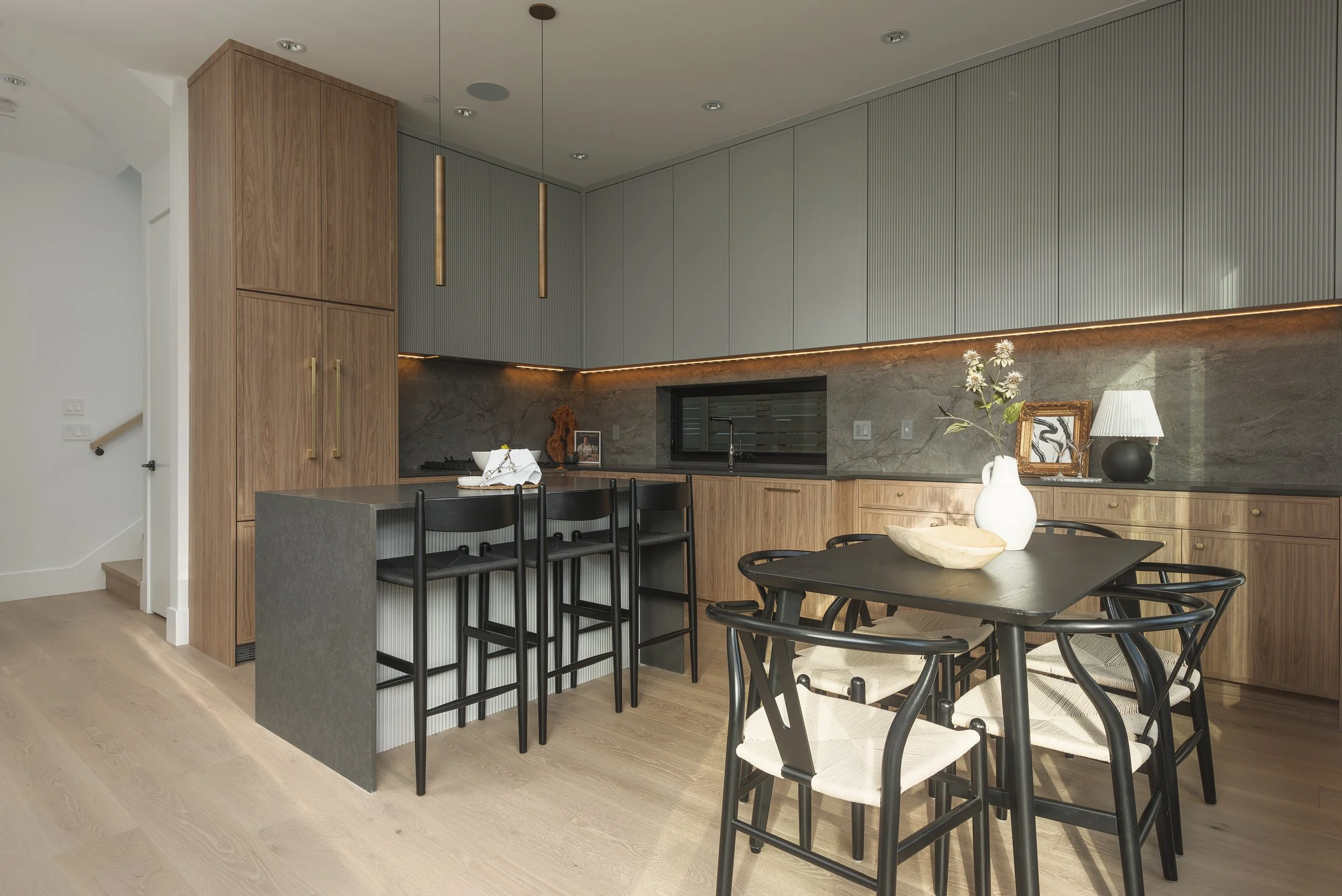 Modern kitchen with wooden cabinets, a black dining table with six chairs, a marble backsplash, and decorative items on the countertop including a vase with flowers, a framed picture, and a lamp.