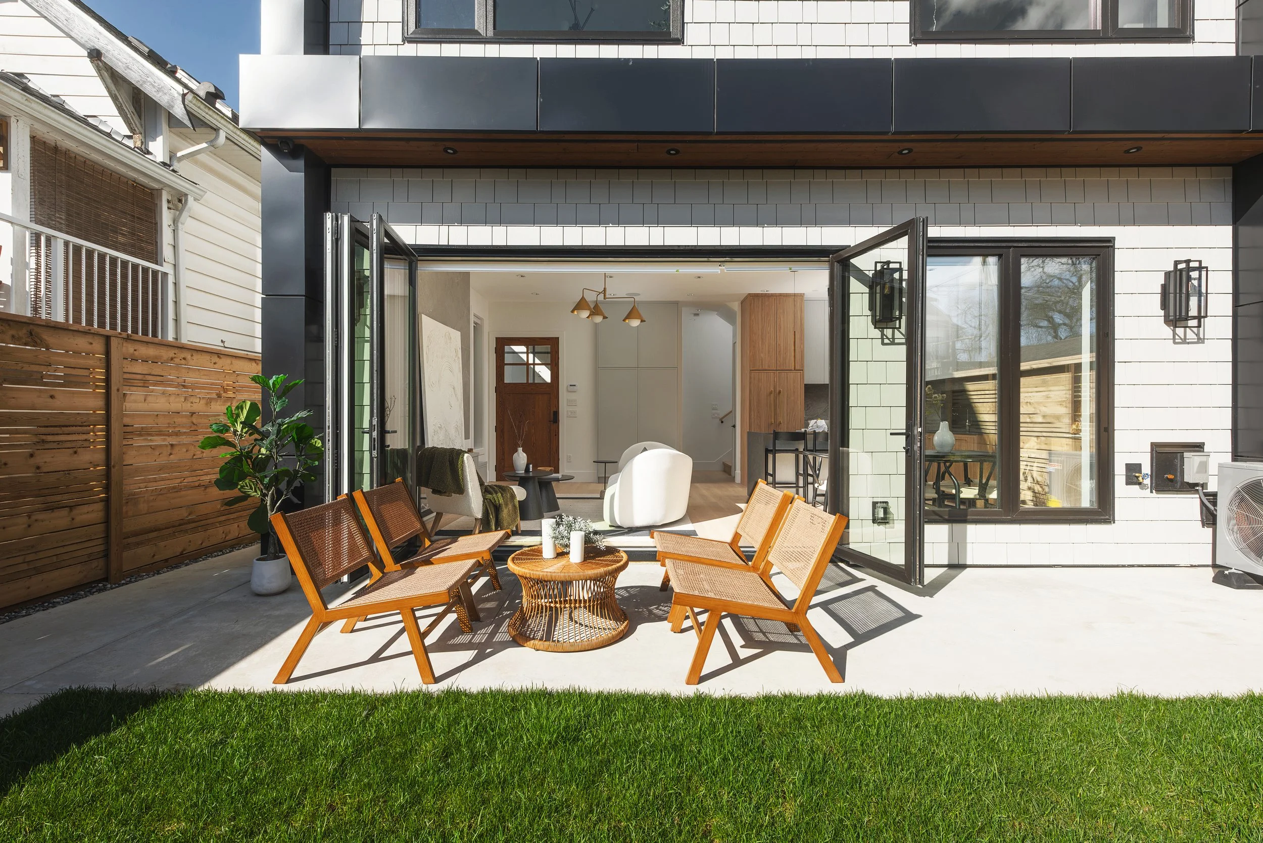 Modern backyard patio with wooden chairs, round wicker table, and open glass doors leading to a bright living room with white and wooden decor, surrounded by a green lawn and a wooden fence.
