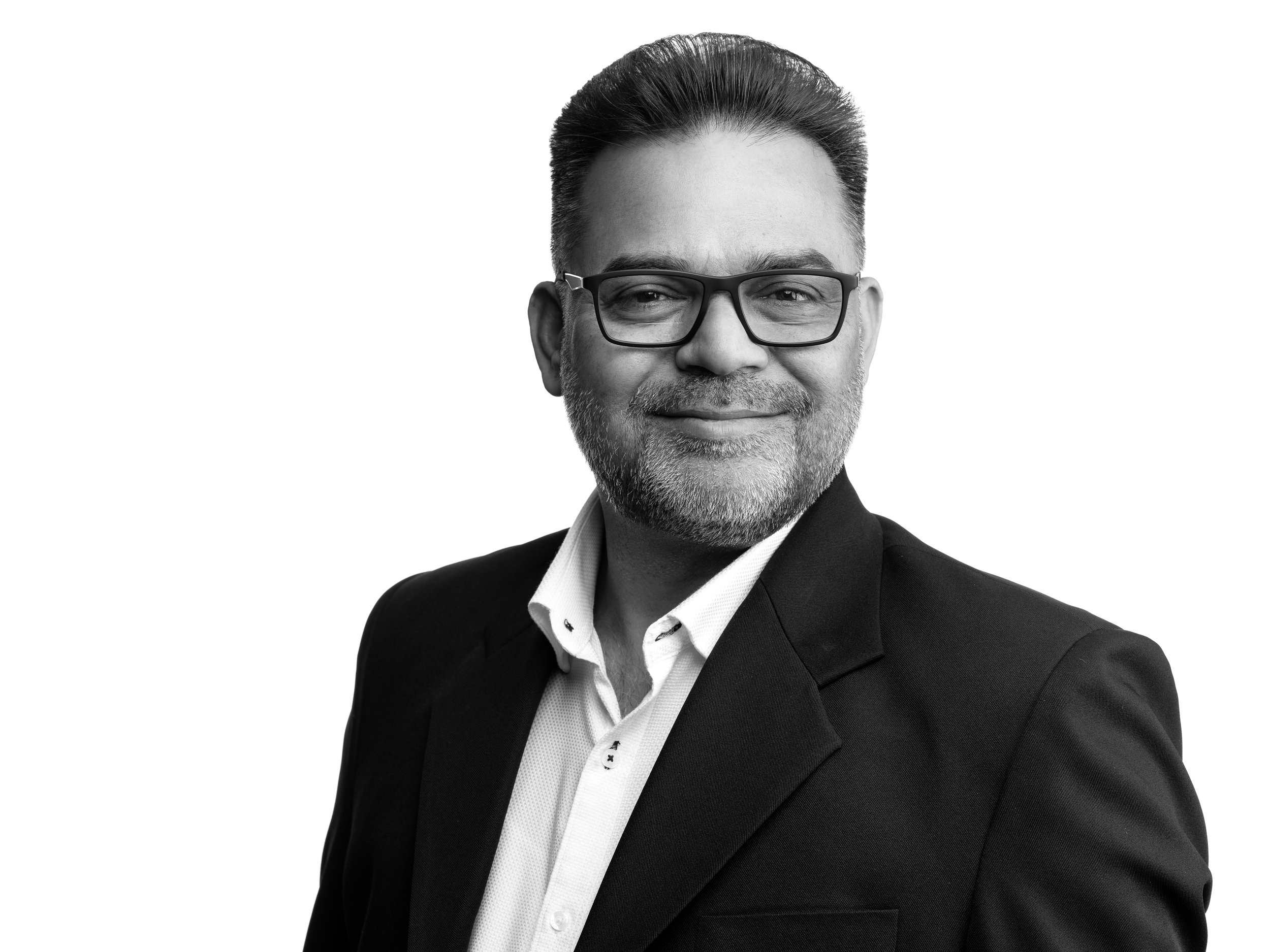 A black and white professional portrait of a middle-aged man with glasses, short dark hair, and a beard, wearing a suit and white shirt, smiling with a confident expression.