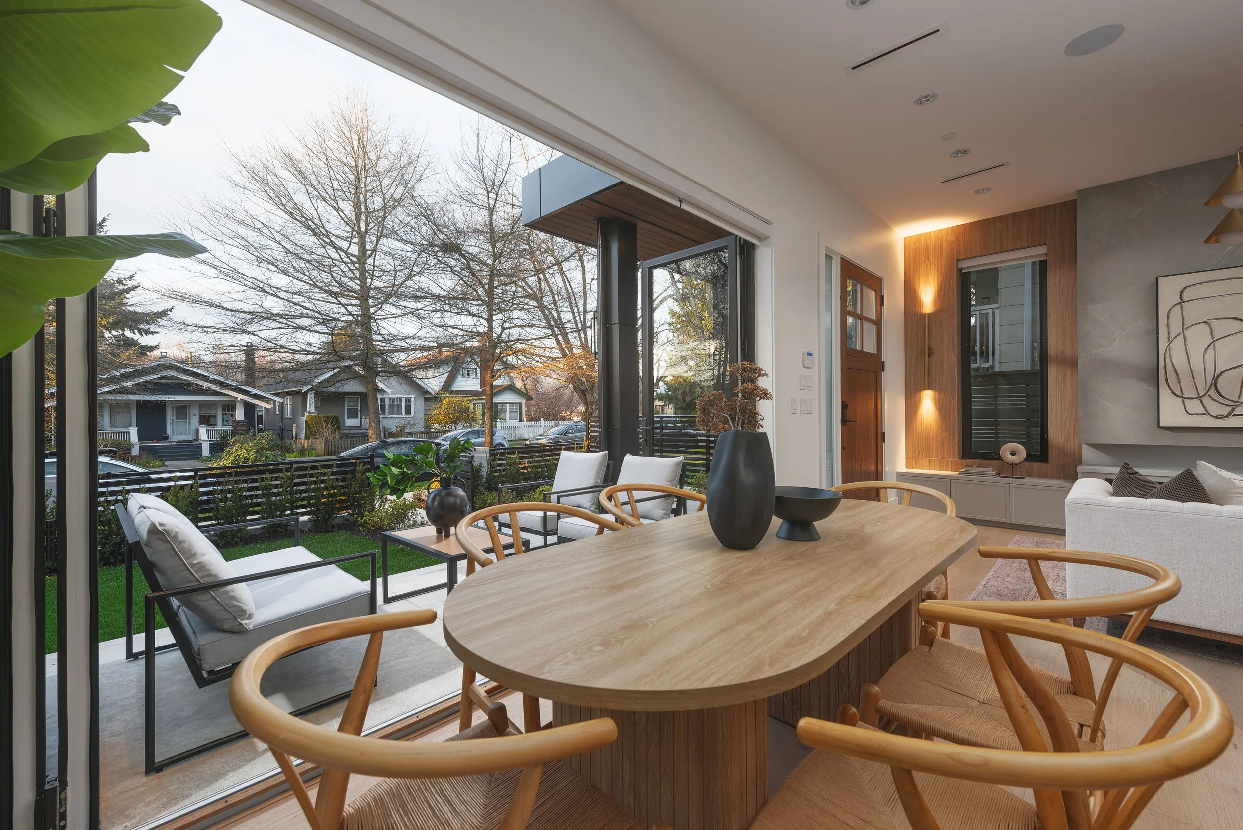 Modern living and dining room with large glass sliding door leading to outdoor patio, featuring wooden dining table with chairs, and white sofa with abstract wall art.