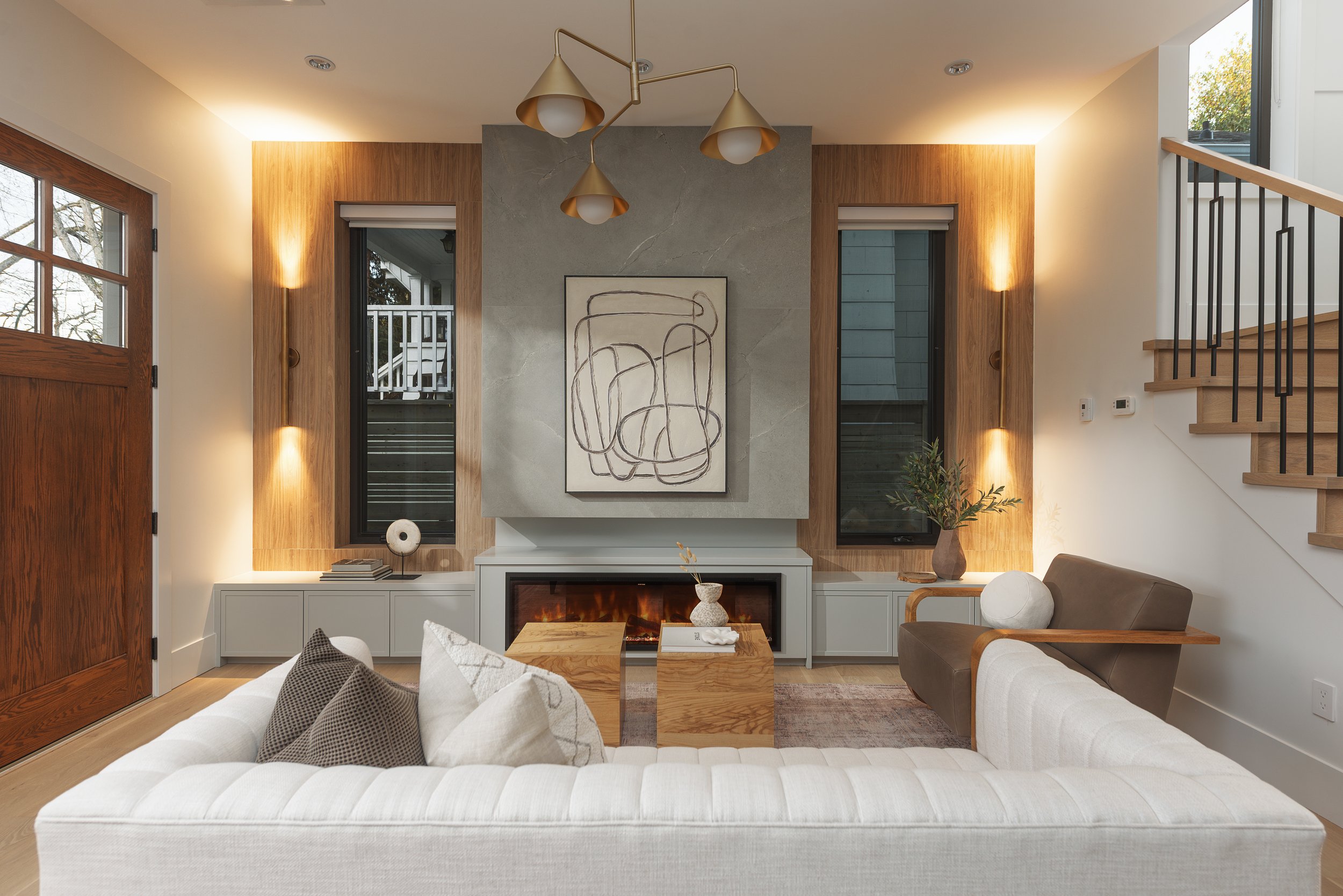 Living room with white sofa, brown armchair, wooden side tables, modern fireplace, abstract art above fireplace, large windows, staircase, and decorative lighting.
